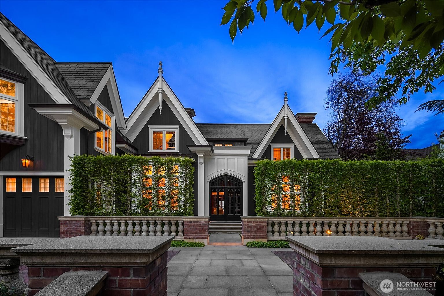 This striking Tudor-style home features a dramatic dark exterior with sharp gabled rooflines and a prominent, arched entryway. A stone pathway leads through a brick-walled courtyard adorned with manicured hedges, creating a sophisticated and private curb appeal. The warm glow from the interior windows contrasts beautifully against the deep blue twilight sky, highlighting the home's architectural elegance.