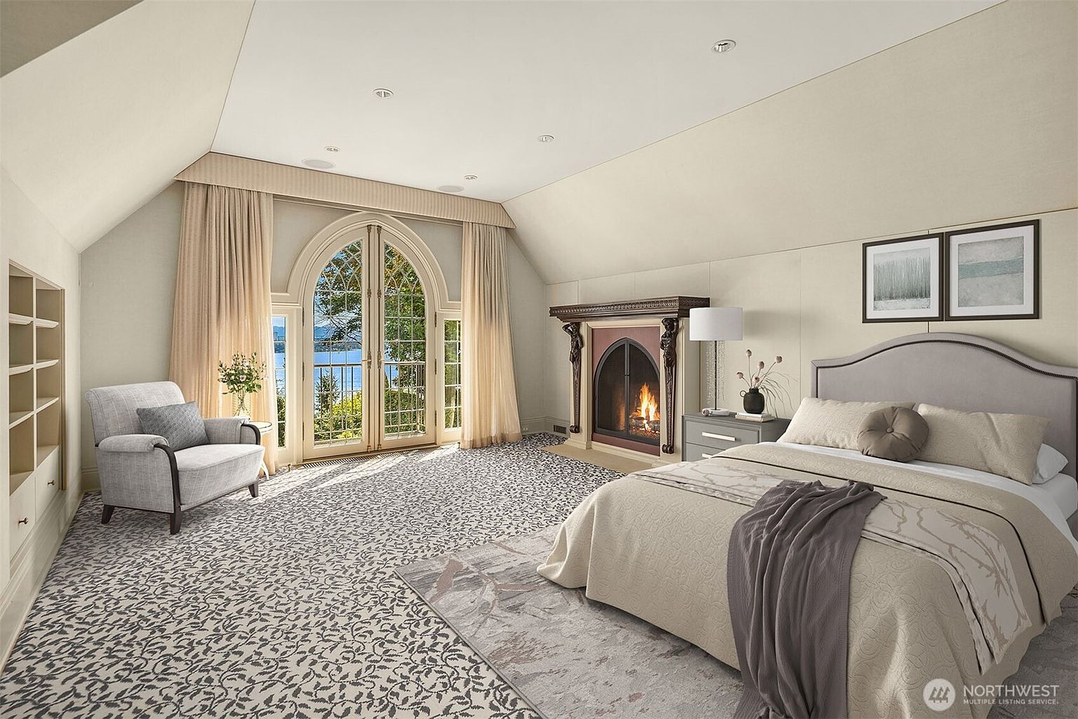 This elegant primary bedroom features a sophisticated neutral color palette, a plush upholstered bed, and a cozy fireplace with a decorative mantel. Large arched French doors provide a stunning view of the water, while patterned carpeting and built-in shelving add texture and character to the spacious, light-filled room.