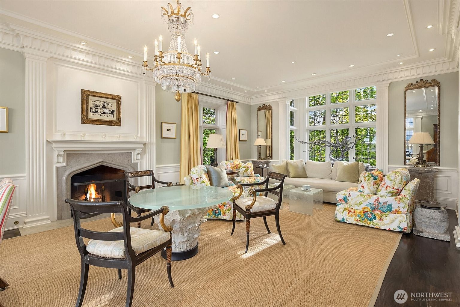 This elegant and spacious living room features a grand crystal chandelier, a classic stone fireplace, and large windows that flood the space with natural light. The room is furnished with a mix of sophisticated floral-patterned armchairs, a neutral sofa, and a unique glass-topped pedestal table, creating a refined yet comfortable atmosphere. The perspective is a wide-angle shot from the corner of the room, showcasing the high ceilings, intricate crown molding, and the seamless blend of traditional architectural details with upscale interior design.