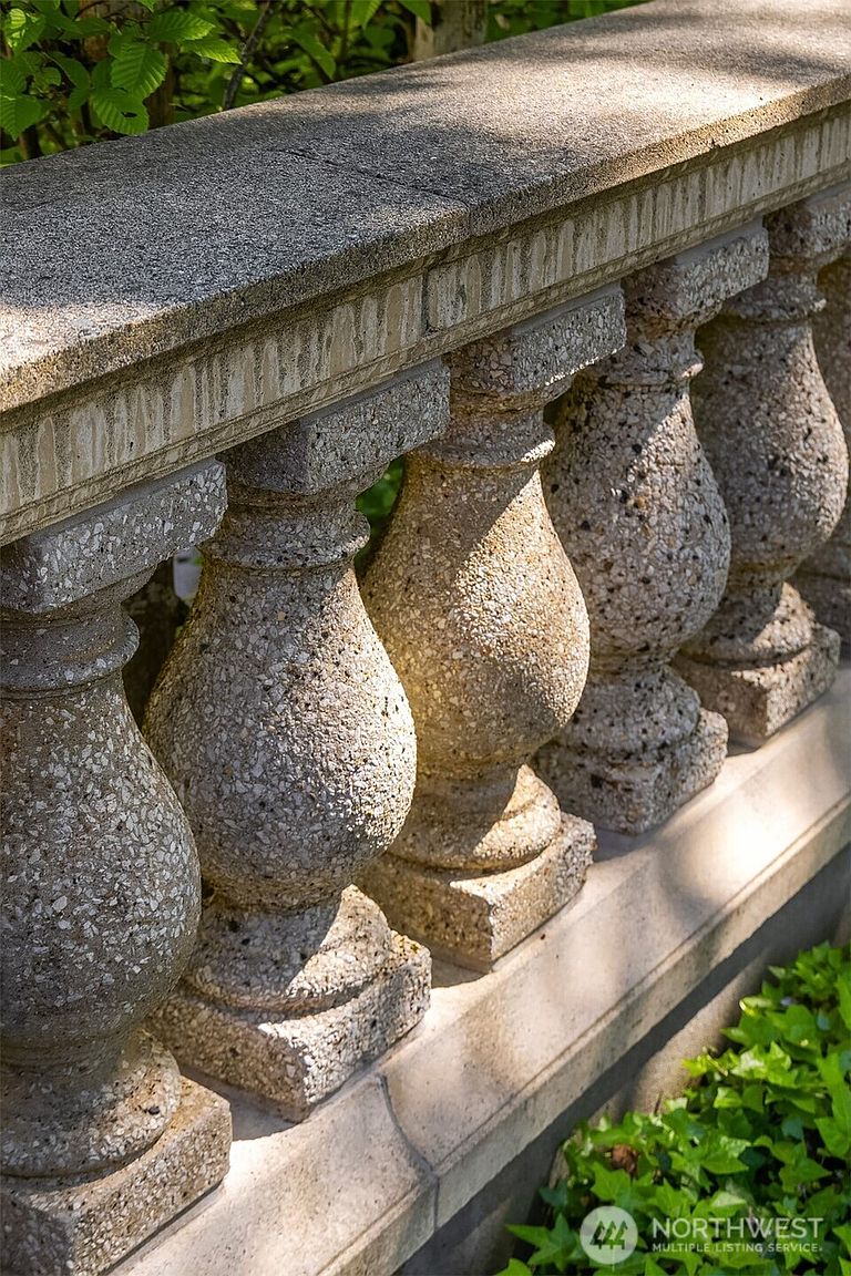 This close-up shot captures a series of weathered, aggregate concrete balusters supporting a heavy stone railing. The perspective is angled along the balustrade, emphasizing the textured, speckled surface of the stone and the play of sunlight and shadow across the architectural elements. Lush green foliage is visible in the background and foreground, suggesting a garden or terrace setting.