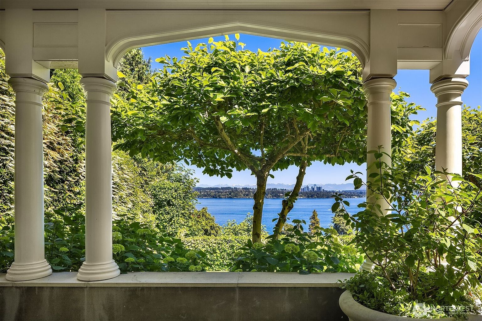 This image captures a serene view from a covered balcony or terrace, framed by elegant white architectural columns. The perspective looks out over a lush, green garden toward a sparkling blue lake with a distant city skyline and mountain range in the background. The scene evokes a sense of tranquility and luxury, highlighting the home's prime waterfront location and scenic vistas.