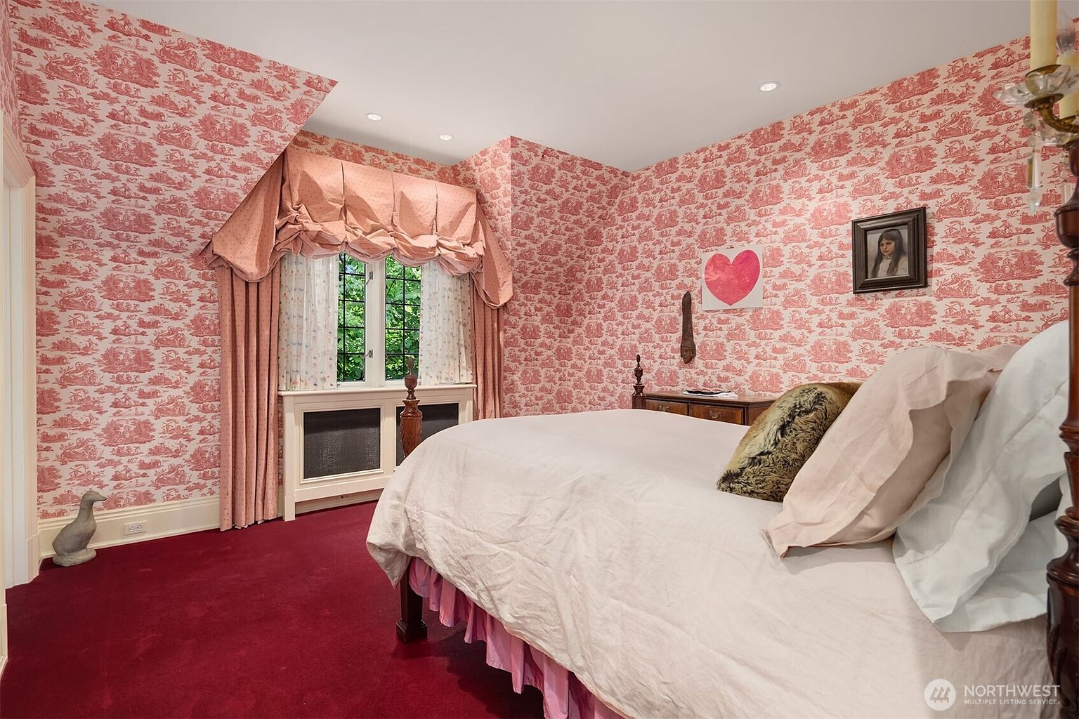 This charming bedroom features distinctive pink toile wallpaper and a matching valance over the window, creating a classic, traditional aesthetic. The room is furnished with a bed dressed in neutral linens, a dark wood dresser, and a framed portrait on the wall, all set against a bold, deep red carpet. The perspective is from the corner of the room, capturing the cozy, vintage-inspired atmosphere.