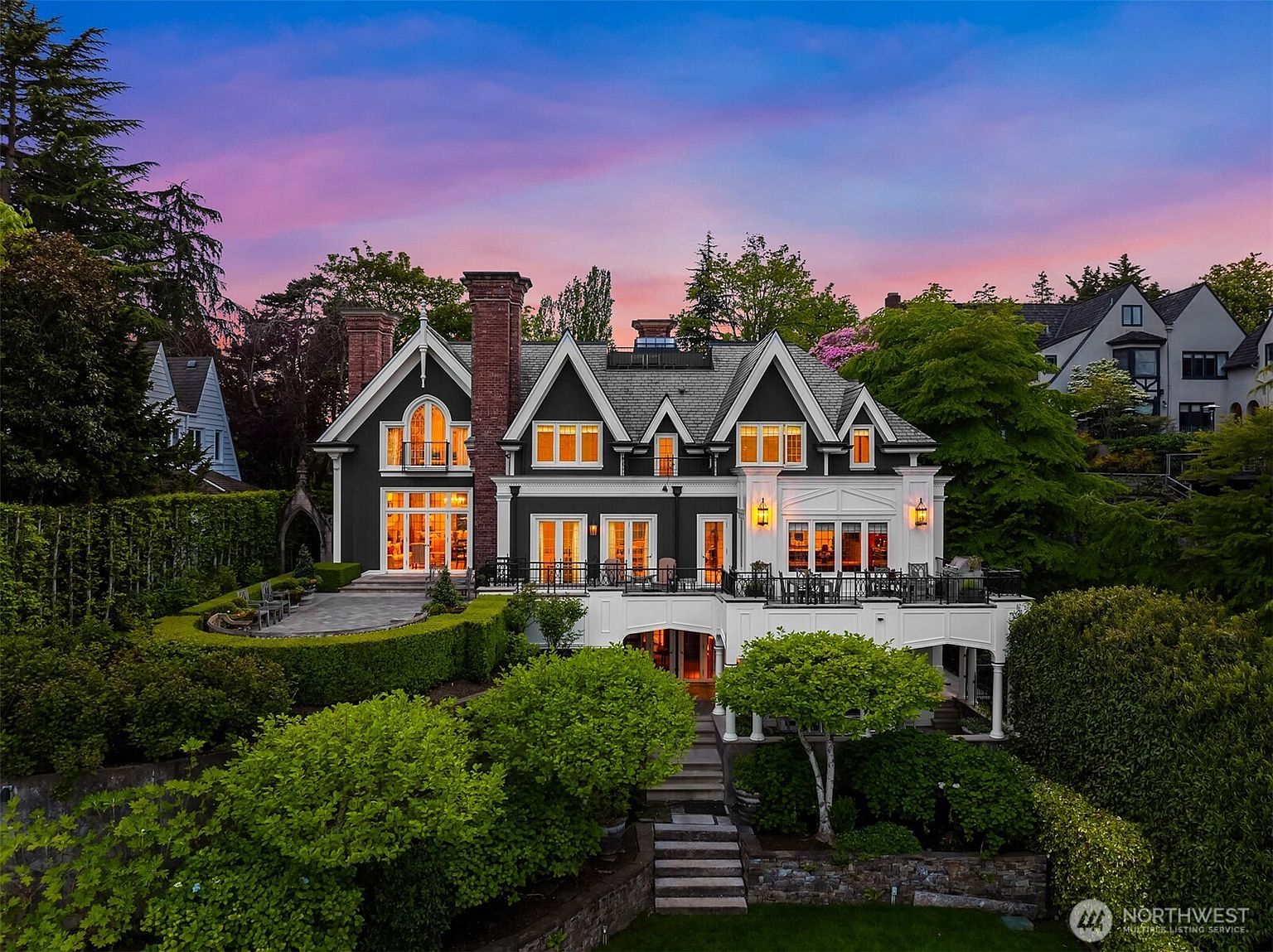 This stunning multi-story estate features a sophisticated Tudor-inspired architectural style with a striking dark grey and white exterior palette. The rear view showcases an expansive elevated terrace, manicured tiered landscaping, and large windows that capture the warm glow of a sunset sky. The property exudes luxury and elegance, perfectly framed by lush greenery and mature trees.
