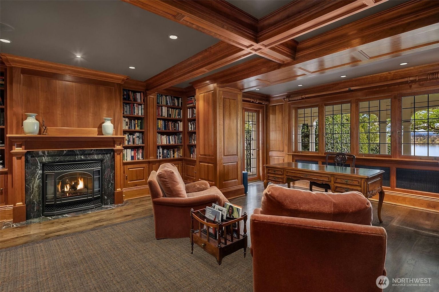 This sophisticated home office features rich, floor-to-ceiling wood paneling and built-in bookshelves, creating a warm and scholarly atmosphere. A grand fireplace with a dark marble surround serves as a focal point, while a classic wooden desk is positioned to overlook the scenic water view through large, multi-paned windows. The room is furnished with plush, velvet-upholstered armchairs, emphasizing a sense of luxury and comfort.
