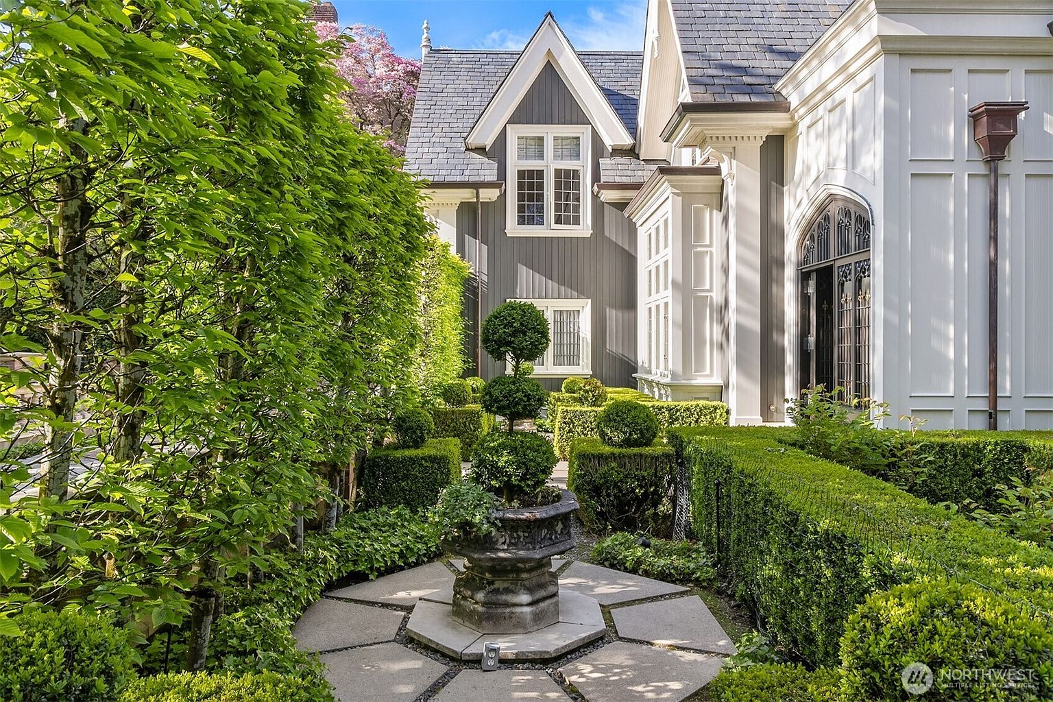 This elegant garden scene features a meticulously manicured landscape with structured boxwood hedges and a central stone planter holding a tiered topiary. The view captures the side of a sophisticated home with a blend of dark grey vertical siding and white architectural trim, framed by lush green foliage and a blooming pink tree in the background. The perspective is eye-level, emphasizing the formal, European-inspired garden design and the refined aesthetic of the property.