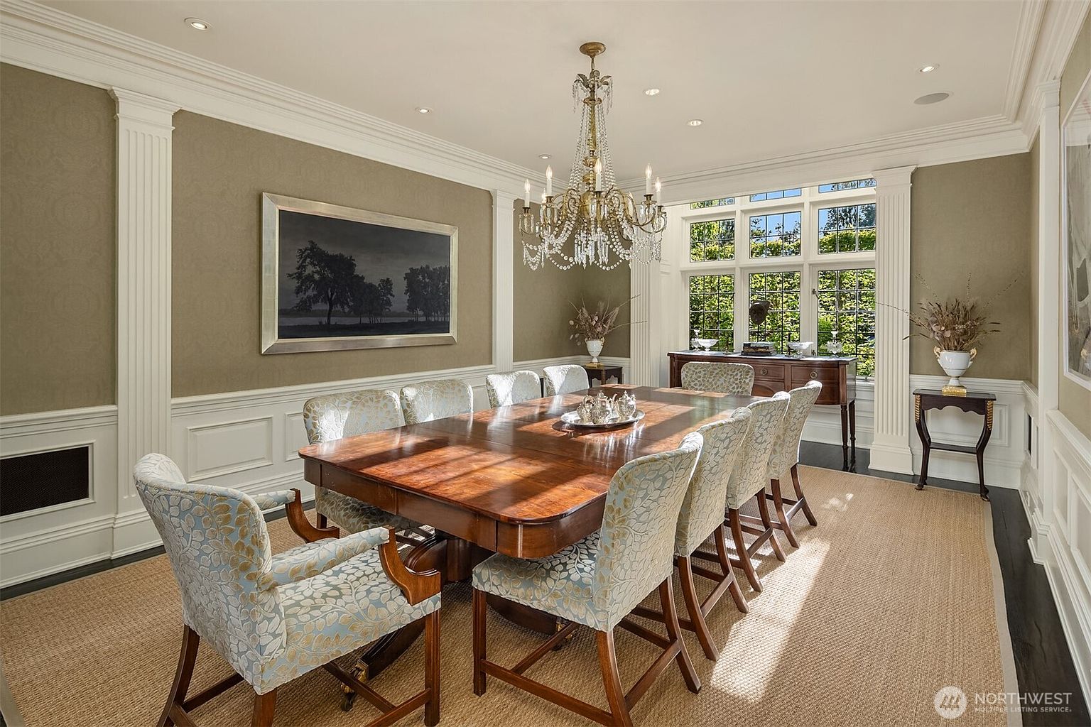 This elegant formal dining room features a large, polished wooden table surrounded by upholstered chairs with a light blue patterned fabric. The room is anchored by a grand crystal chandelier, wainscoting along the walls, and a large window that allows natural light to illuminate the space. A sideboard and a small accent table provide additional storage and display space, creating a sophisticated and inviting atmosphere for formal gatherings.