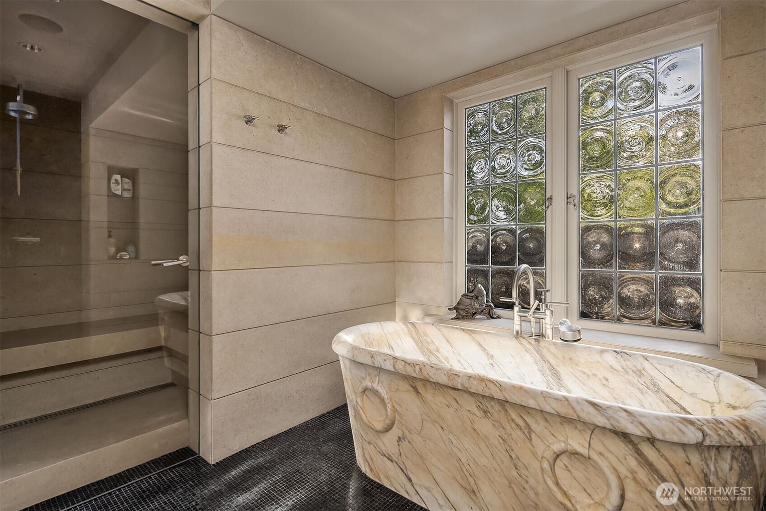 This luxurious primary bathroom features a stunning, custom-carved marble soaking tub as its centerpiece, positioned beneath a unique glass block window. To the left, a spacious walk-in shower with a glass door and stone-tiled walls adds to the spa-like atmosphere, while the dark mosaic floor tiles provide a sophisticated contrast. The overall aesthetic is one of high-end elegance and serene relaxation.