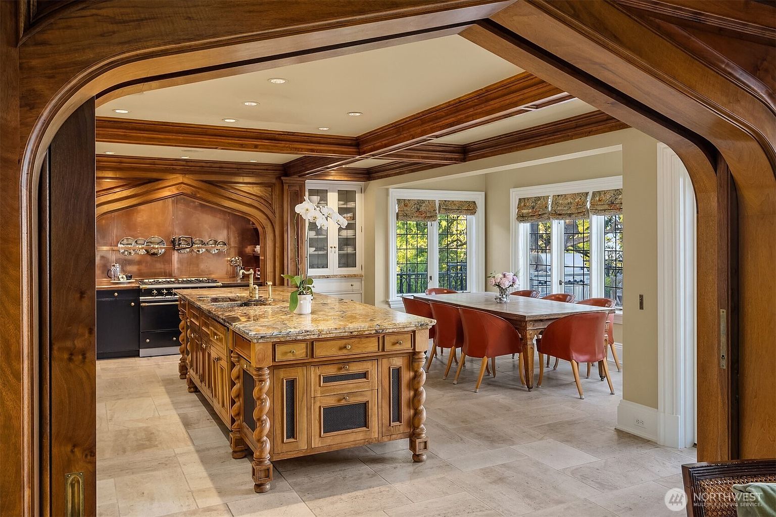 This elegant, traditional-style kitchen features a large, ornate wooden island with a granite countertop and a copper-backsplash cooking area. The space is framed by rich, dark wood trim and coffered ceilings, transitioning seamlessly into a formal dining area with large windows. The overall impression is one of sophisticated craftsmanship and warm, inviting luxury.