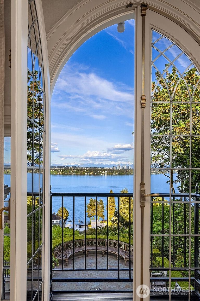 This image captures a stunning, elevated view from an arched doorway leading onto a private balcony overlooking a serene lake and distant mountain range. The balcony features elegant black wrought-iron railings and a curved bench, creating a sophisticated outdoor retreat. The perspective frames the picturesque landscape through the architectural detail of the French doors, emphasizing a seamless connection between indoor luxury and natural beauty.