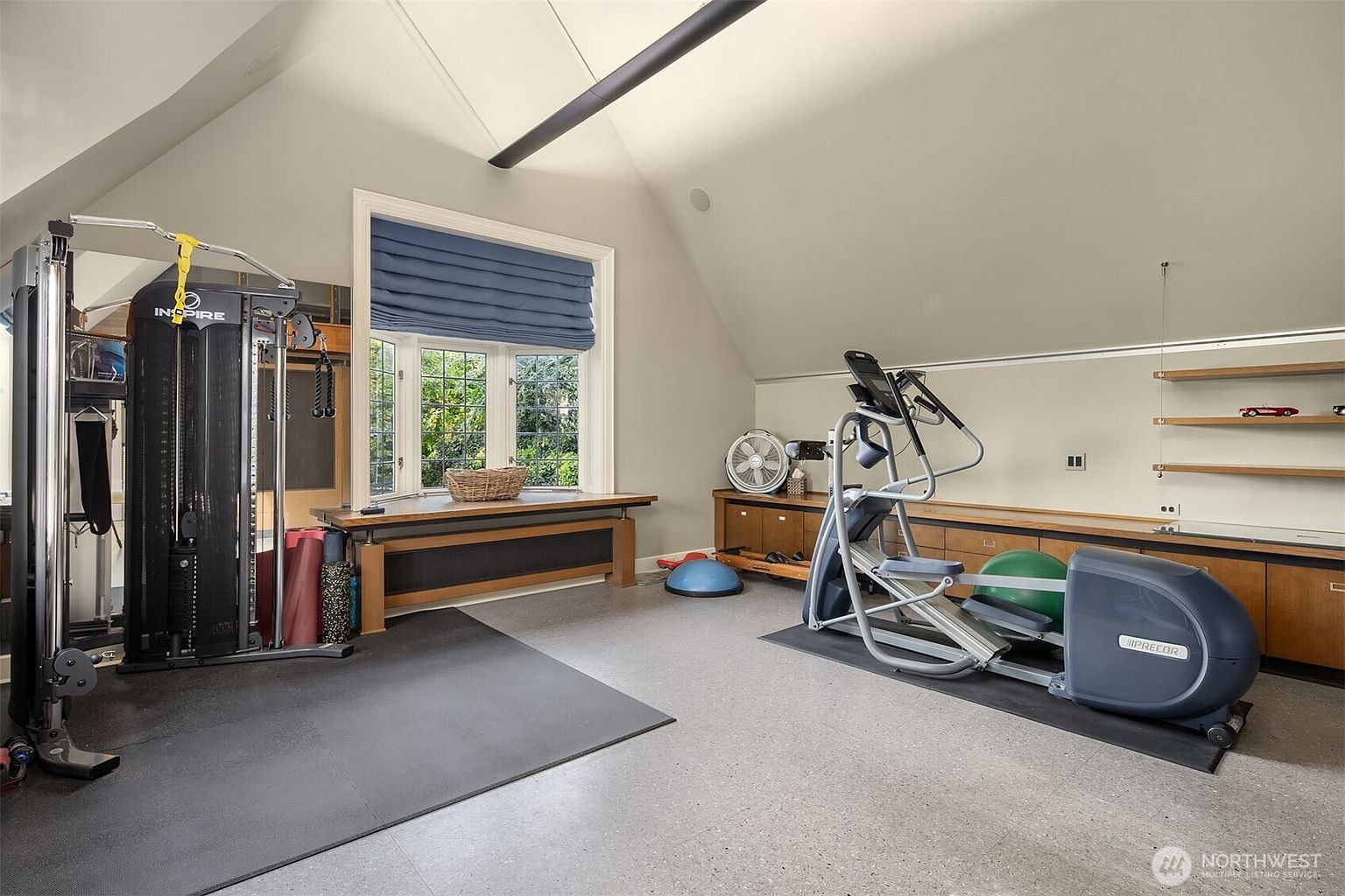 This well-equipped home gym features a vaulted ceiling and a large bay window that brings in natural light, creating an airy and inviting workout space. The room is furnished with a professional-grade Inspire functional trainer, a Precor elliptical machine, and various fitness accessories like a Bosu ball and yoga mats. The space is finished with durable flooring, built-in wooden cabinetry for storage, and floating shelves, offering a functional and organized environment for fitness enthusiasts.