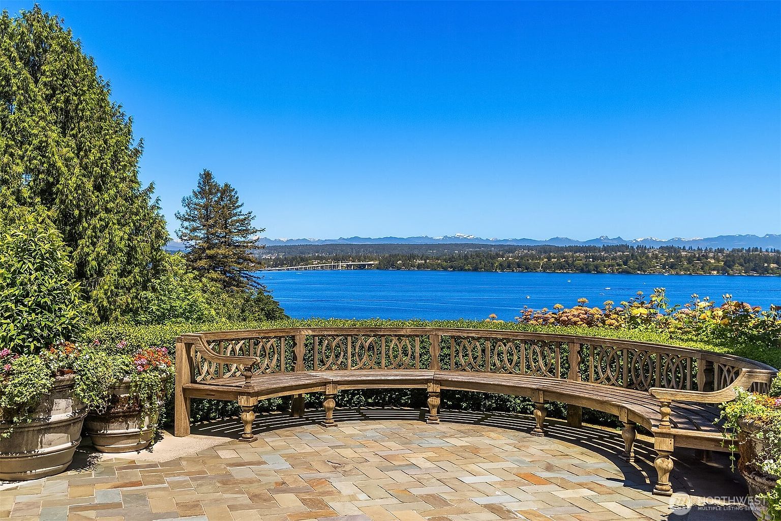 This scenic outdoor patio features a curved, ornate wooden bench set against a backdrop of a stunning lake view and distant mountain peaks. The stone-paved terrace is framed by lush greenery and potted plants, creating a serene and elegant atmosphere perfect for relaxation. The perspective captures the expansive horizon, emphasizing the property's prime waterfront location and tranquil setting.