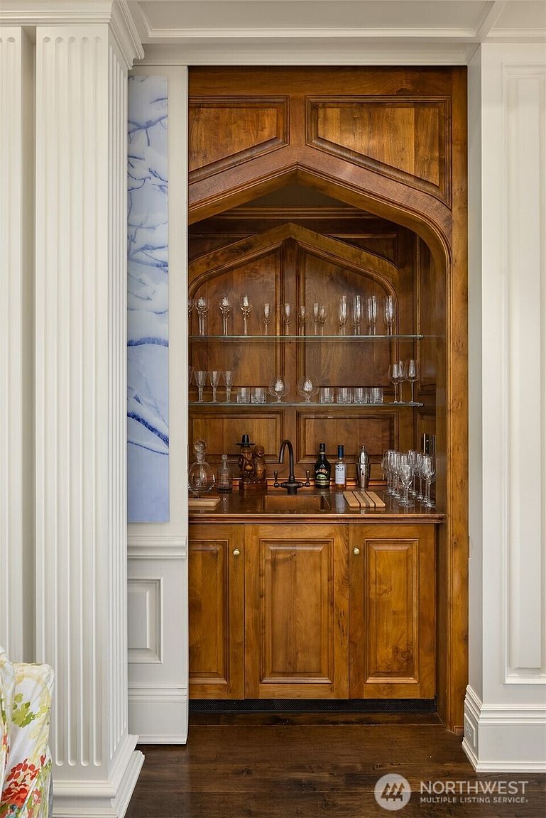This image features a sophisticated, built-in wet bar nestled within a custom-crafted wooden alcove with an elegant arched design. The space is equipped with a small sink, glass shelving displaying a collection of stemware, and rich, warm-toned cabinetry that contrasts beautifully with the surrounding white architectural trim. The perspective is a straight-on, eye-level shot that highlights the craftsmanship and luxurious, classic aesthetic of the home's interior.