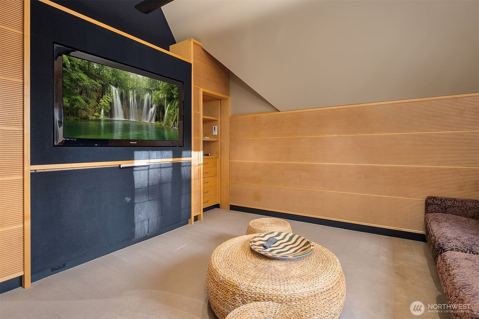 This cozy media room features a large wall-mounted television set against a dark, textured accent wall framed by light wood trim. The room includes a unique sloped ceiling, built-in shelving, and two round, woven ottomans serving as a coffee table in front of a plush sofa. The overall aesthetic is warm and contemporary, creating an inviting space for relaxation or entertainment.