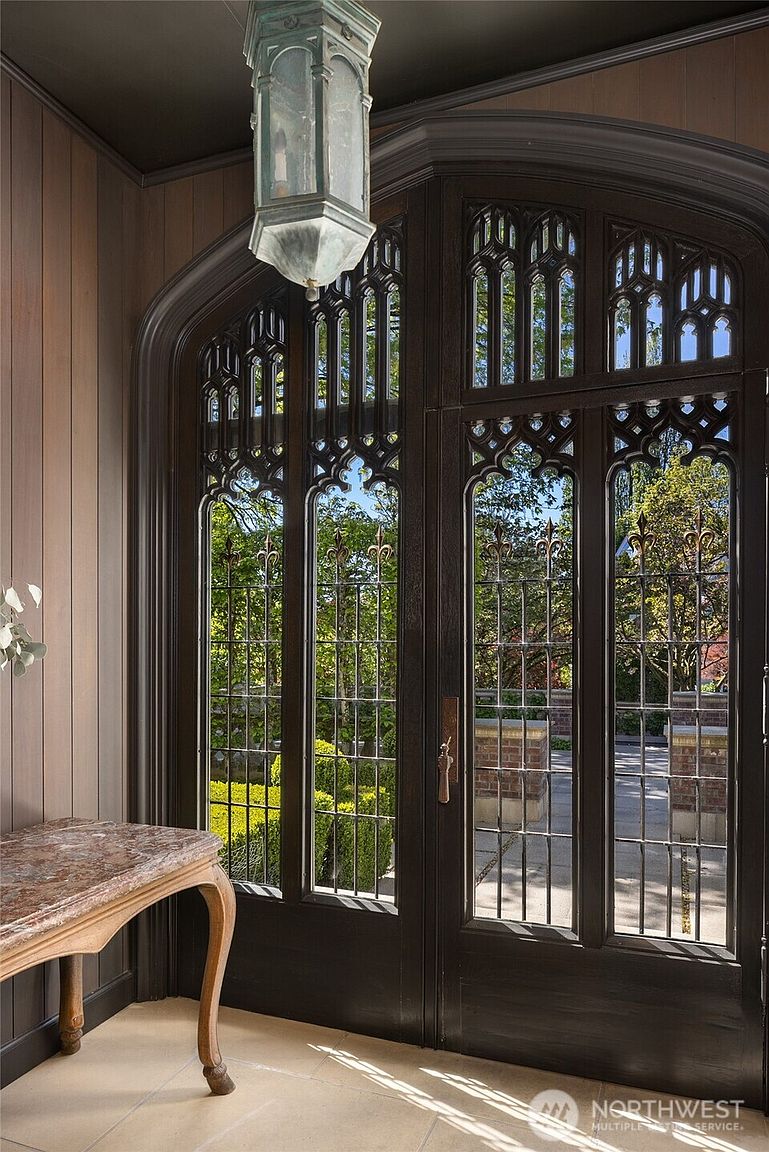 This elegant entryway features a grand, arched double door with intricate Gothic-style woodwork and glass panels that allow natural light to flood the space. A sophisticated marble-topped console table sits to the left, while a vintage-inspired lantern pendant light hangs from the ceiling, adding a touch of historic charm. The perspective is from inside the home, looking out through the ornate entryway toward a manicured garden, creating a welcoming and luxurious first impression.