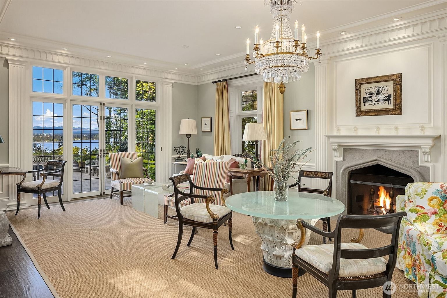 This elegant, light-filled living room features a sophisticated traditional design with a grand crystal chandelier as its centerpiece. The space is anchored by a stone fireplace and large French doors that open to a scenic waterfront view, while a mix of patterned and neutral seating creates a refined yet comfortable atmosphere. The room is finished with crown molding and a textured area rug, offering a luxurious and inviting aesthetic.