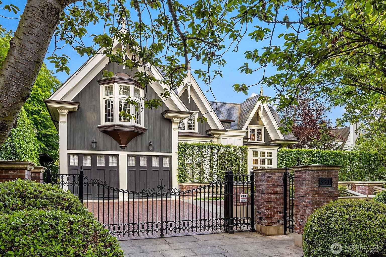 This elegant, Tudor-inspired home features a striking dark grey and white exterior with a prominent bay window and steep, gabled rooflines. A decorative black wrought-iron gate and brick pillars frame the entrance to a brick-paved driveway, creating a sense of privacy and sophistication. The property is surrounded by lush, manicured greenery, contributing to a refined and welcoming curb appeal.