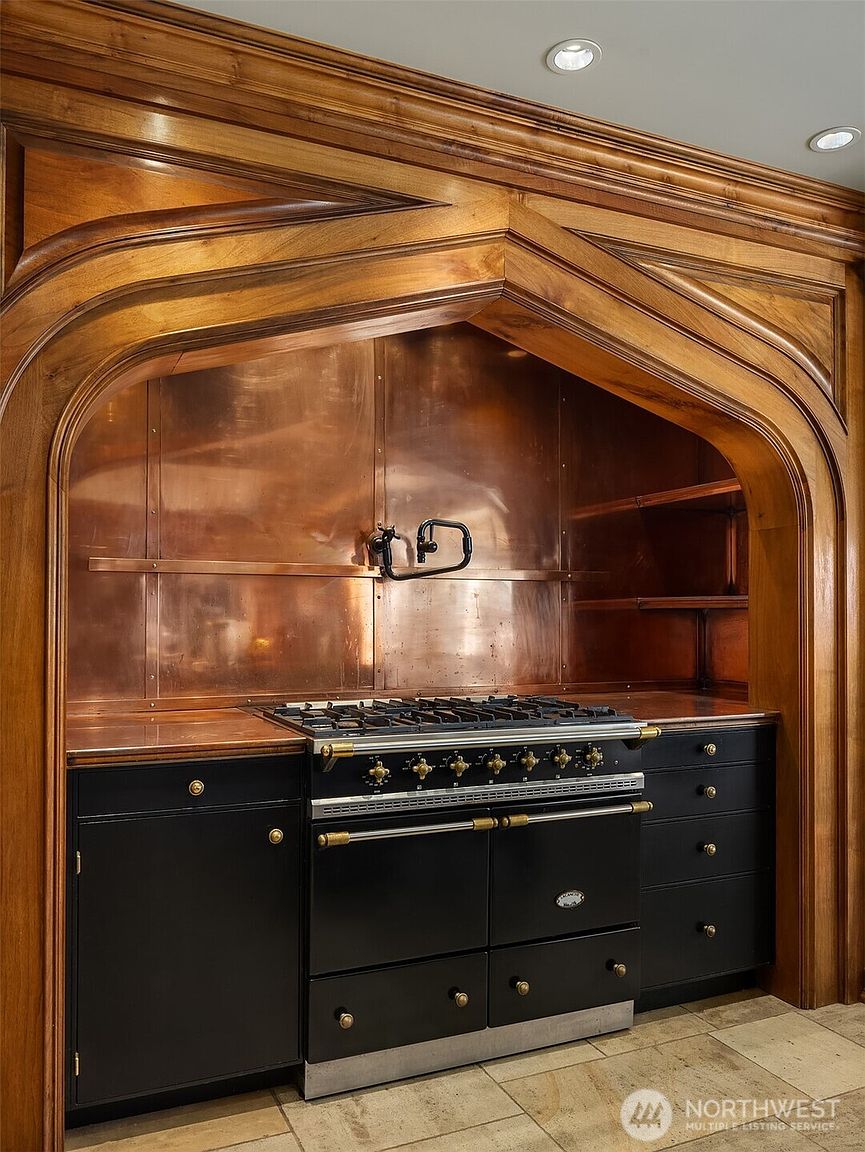 This high-end kitchen features a dramatic, custom-built arched wooden alcove housing a professional-grade black range and a striking copper backsplash. The combination of rich, warm wood tones, dark cabinetry, and metallic accents creates a sophisticated, Old World aesthetic. The perspective is a straight-on, eye-level shot that highlights the craftsmanship of the range hood and the integrated design of the cooking station.