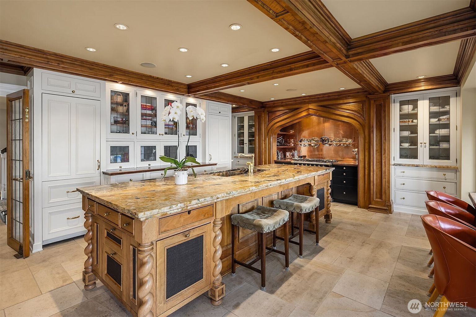 This luxurious, traditional-style kitchen features a massive central island with a rich, patterned granite countertop and intricate wooden base detailing. The space is defined by warm, custom-stained wood cabinetry and ceiling beams, contrasted by elegant white glass-fronted cabinets and a striking copper-hooded stove alcove. The perspective is a wide-angle shot from the dining area, showcasing the sophisticated blend of textures, high-end finishes, and professional-grade appliances.