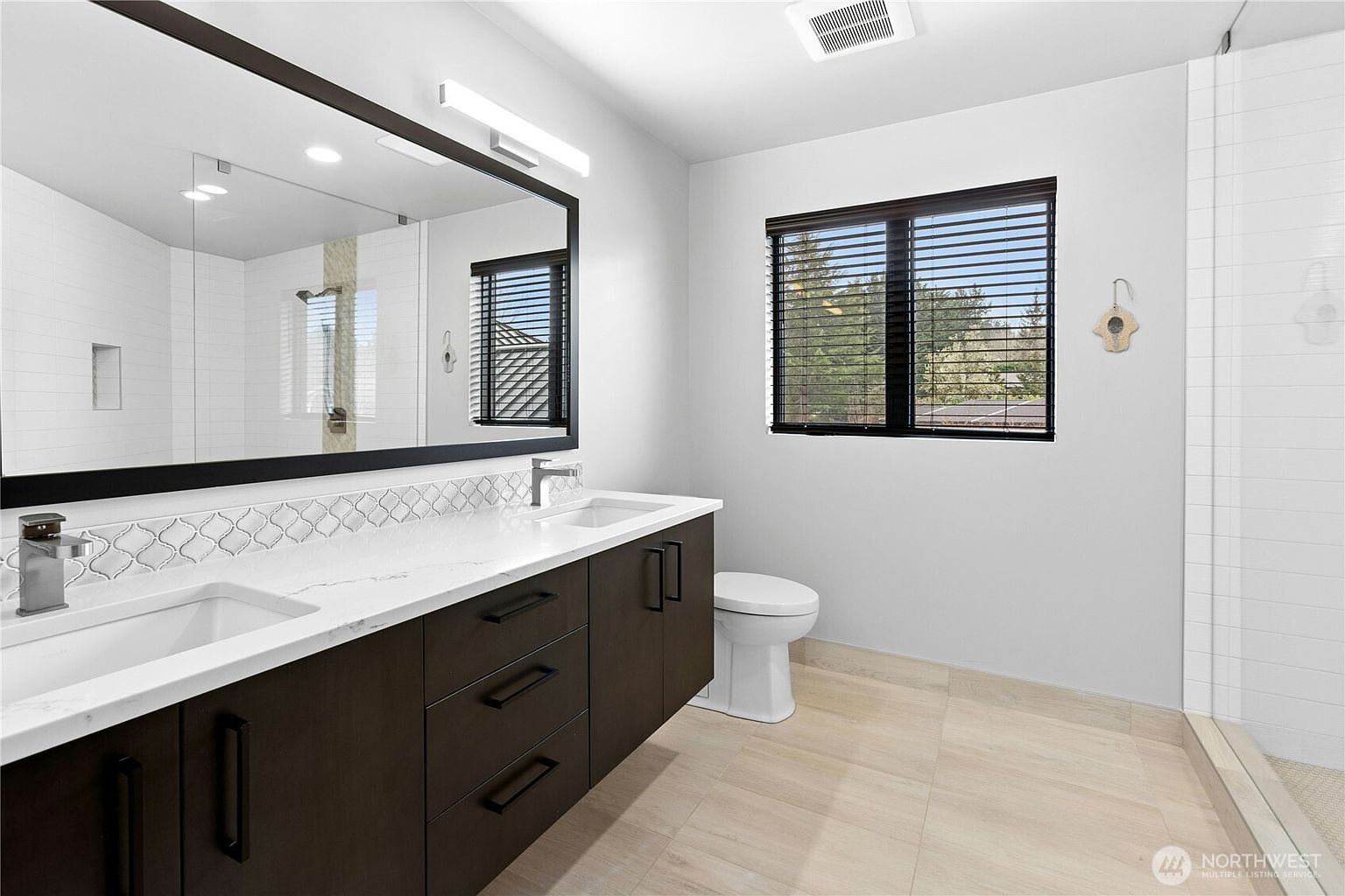 This modern primary bathroom features a sleek double-sink vanity with dark wood cabinetry and a white quartz countertop. A large, expansive mirror spans the wall above the vanity, reflecting the walk-in shower area, while light-colored tile flooring and a window with dark blinds create a bright, contemporary aesthetic. The space is clean and well-lit, offering a sophisticated and functional design.