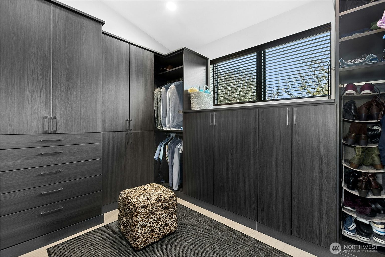 This sophisticated walk-in closet features dark, wood-grain cabinetry that provides ample storage space, including hanging racks, drawers, and open shelving. A central leopard-print ottoman sits on a dark area rug, adding a touch of personality to the sleek, modern design. A window with horizontal blinds allows natural light to brighten the space, while a dedicated shoe rack keeps footwear organized.
