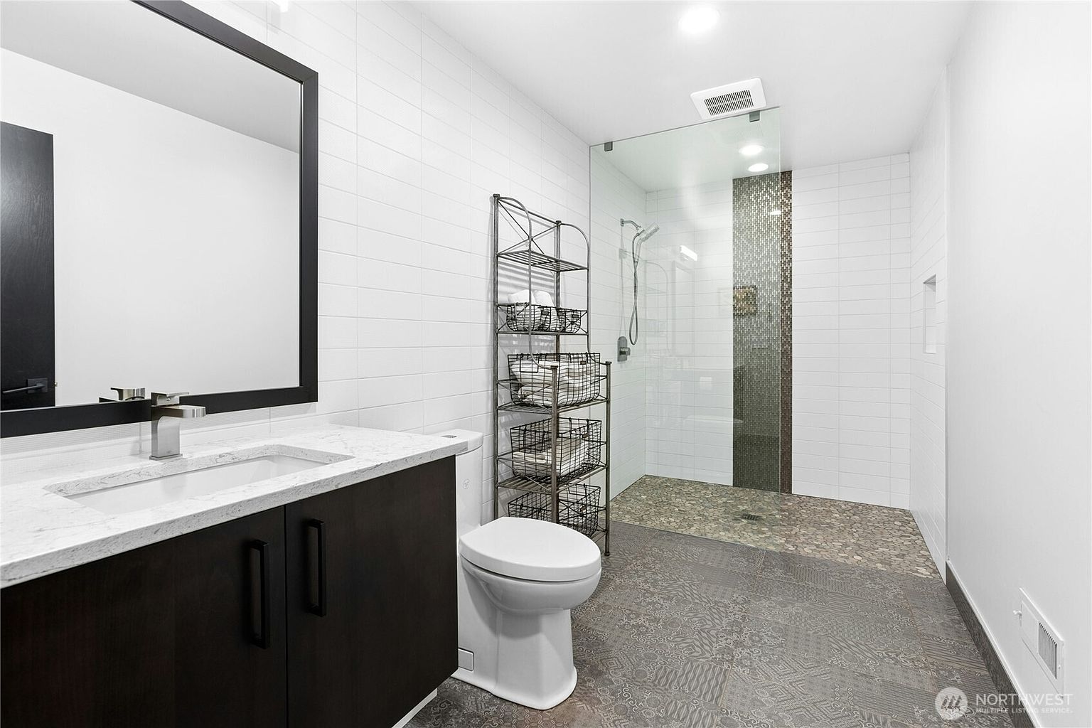 This modern bathroom features a sleek vanity with a white stone countertop, a dark wood cabinet, and a large rectangular mirror. The space includes a walk-in shower with a pebble-stone floor and a decorative mosaic tile accent wall, complemented by a tall metal shelving unit for storage. The overall aesthetic is contemporary and clean, utilizing a neutral color palette to create a spa-like atmosphere.