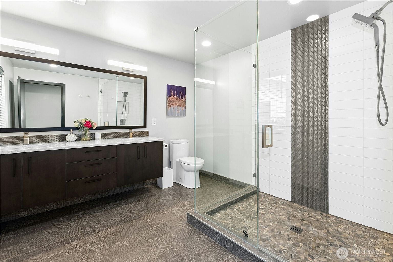 This modern primary bathroom features a sleek, dark wood double vanity with a white countertop and a large, framed mirror. To the right, a spacious walk-in shower is enclosed by glass and showcases a pebble-stone floor and a decorative vertical mosaic tile accent wall. The room is finished with patterned floor tiles, creating a contemporary and sophisticated aesthetic.