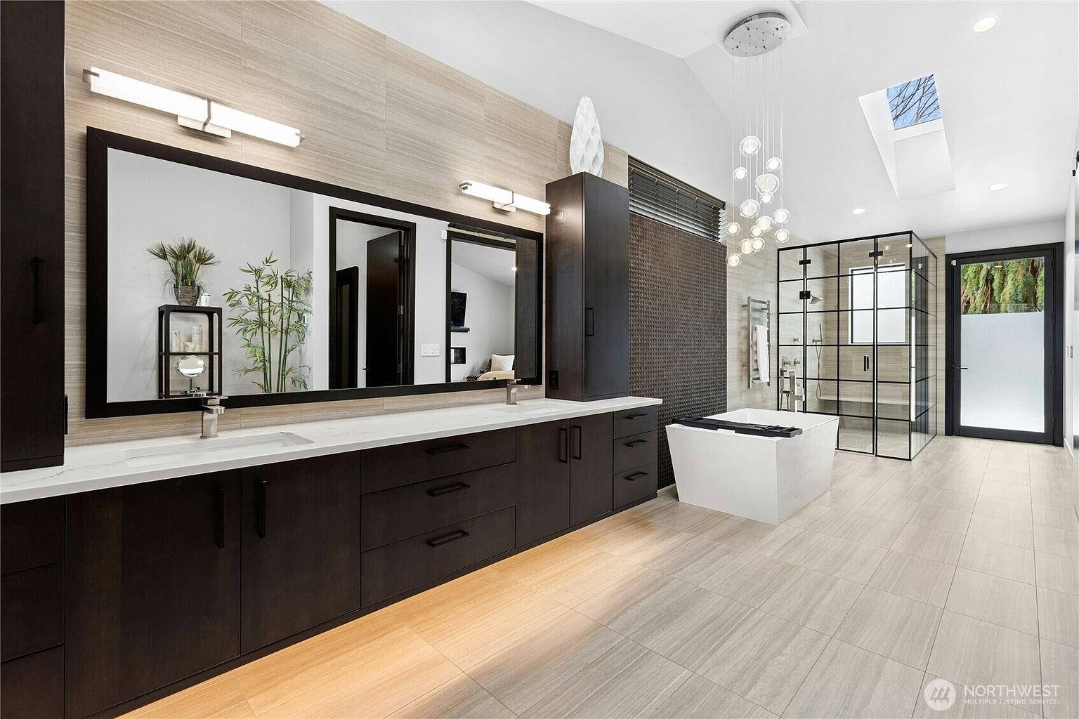 This luxurious primary bathroom features a sleek, modern design with a long dark wood vanity, dual sinks, and a large wall-to-wall mirror. The space is highlighted by a freestanding soaking tub, a spacious walk-in glass shower with black framing, and elegant pendant lighting hanging from the vaulted ceiling. The room is finished with neutral large-format floor tiles and a skylight that fills the area with natural light, creating a sophisticated and spa-like atmosphere.