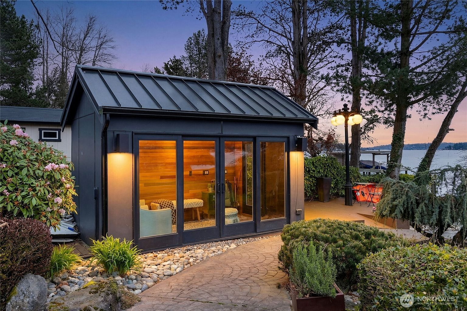 This image showcases a modern, dark-toned detached studio or sauna structure nestled in a lush, landscaped garden setting at dusk. The building features floor-to-ceiling glass doors that reveal a warm, wood-paneled interior, complemented by a stone pathway and a wooden deck overlooking a serene lake. The cinematic perspective captures the inviting glow of the structure against the twilight sky, emphasizing a peaceful and luxurious outdoor retreat.