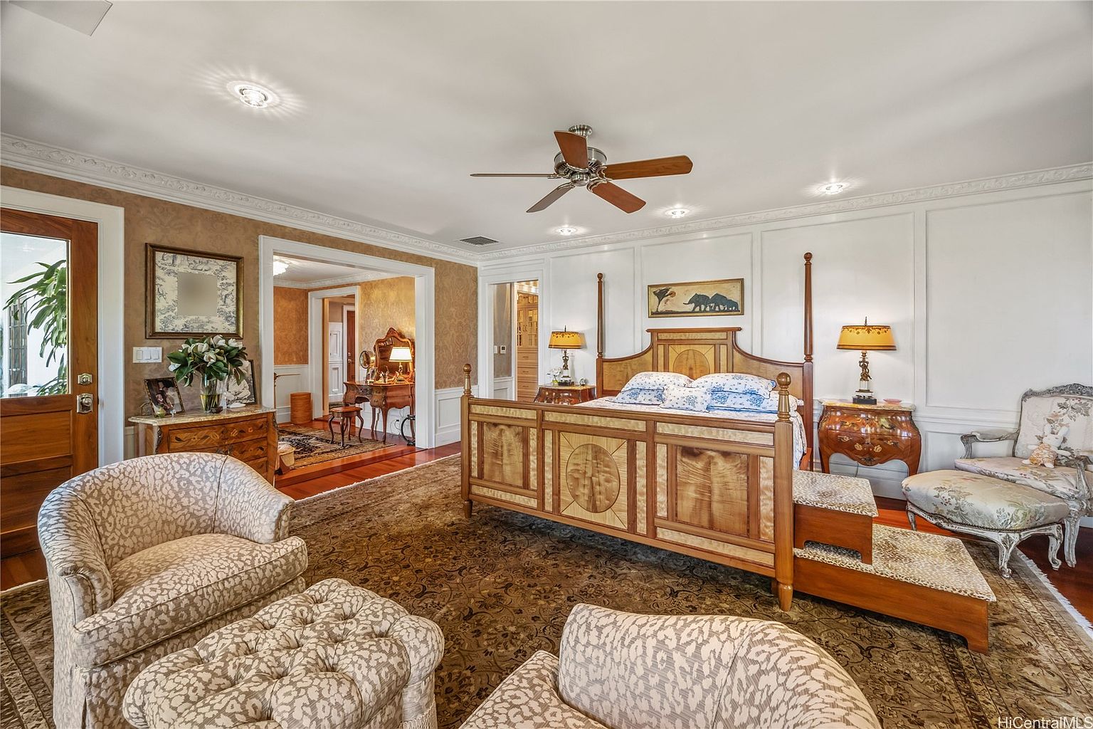 This is a primary bedroom featuring a four-poster bed with a wooden frame and patterned bedding. The room is decorated with a mix of textures and patterns, including a large area rug, upholstered chairs, and decorative lamps. The space has a luxurious and comfortable feel, with an adjacent room visible through an open doorway.