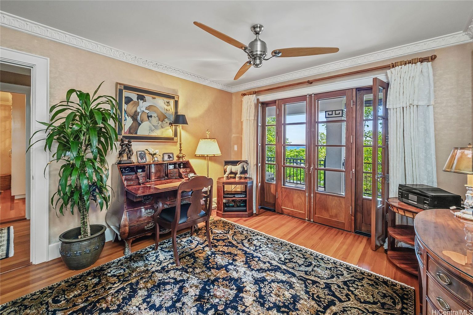 This is an interior shot of a home office or study. The room features a wooden desk, a chair, and a large area rug with a floral pattern. French doors lead to an outdoor balcony, and a ceiling fan is visible above. The room has a warm and inviting atmosphere, perfect for a home office.