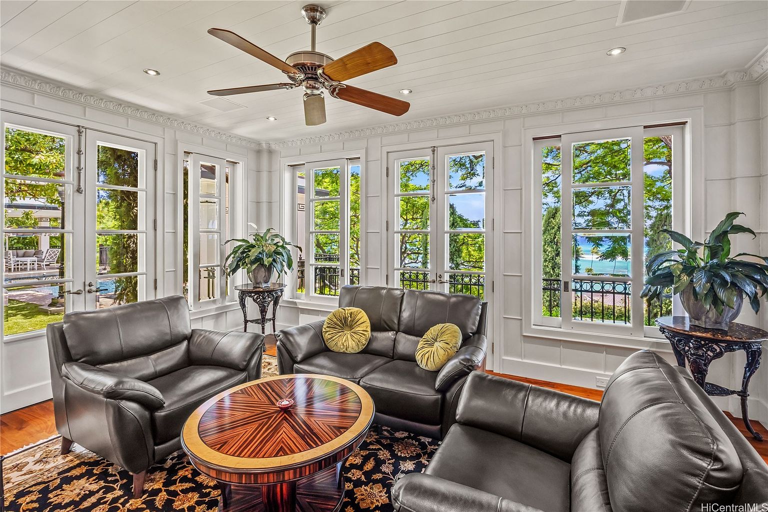This is an interior shot of a sunroom or living room featuring a seating area with dark leather armchairs and a loveseat arranged around a round wooden coffee table. Large windows offer views of the exterior landscape, including trees and a glimpse of the ocean. The room is decorated in a classic style with white paneled walls and a decorative ceiling fan.