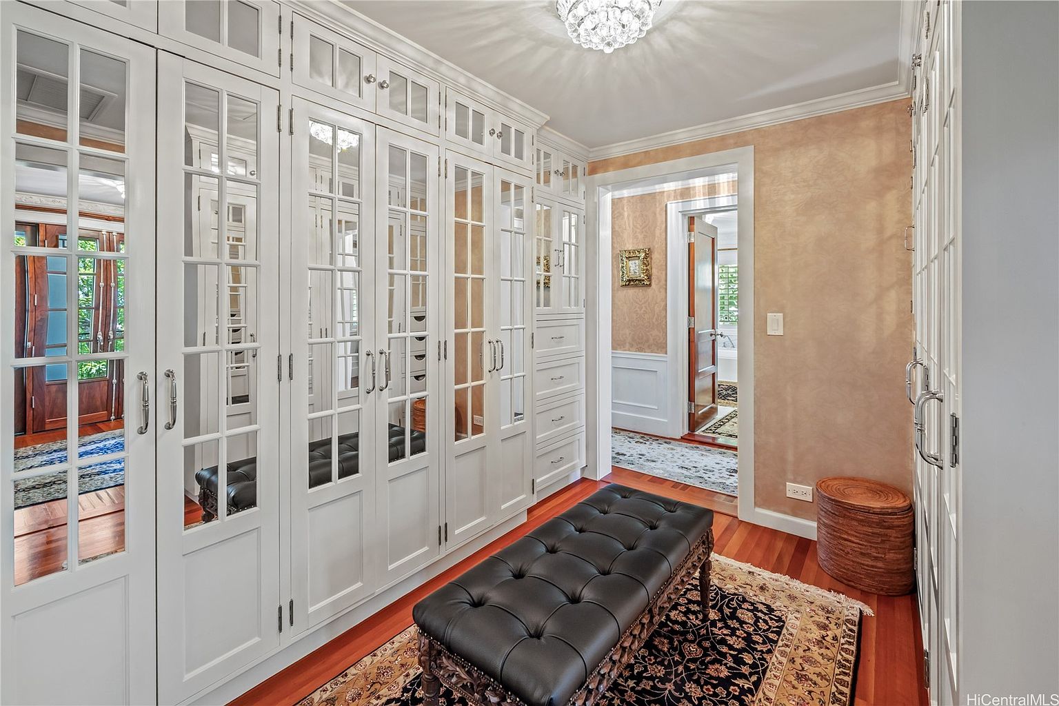 This is a well-organized walk-in closet featuring custom white cabinetry with glass-paneled doors and drawers. A tufted black bench sits on a patterned rug, adding a touch of luxury. The closet is illuminated by a decorative chandelier, and an open doorway leads to another room, suggesting ample space and connectivity within the home.