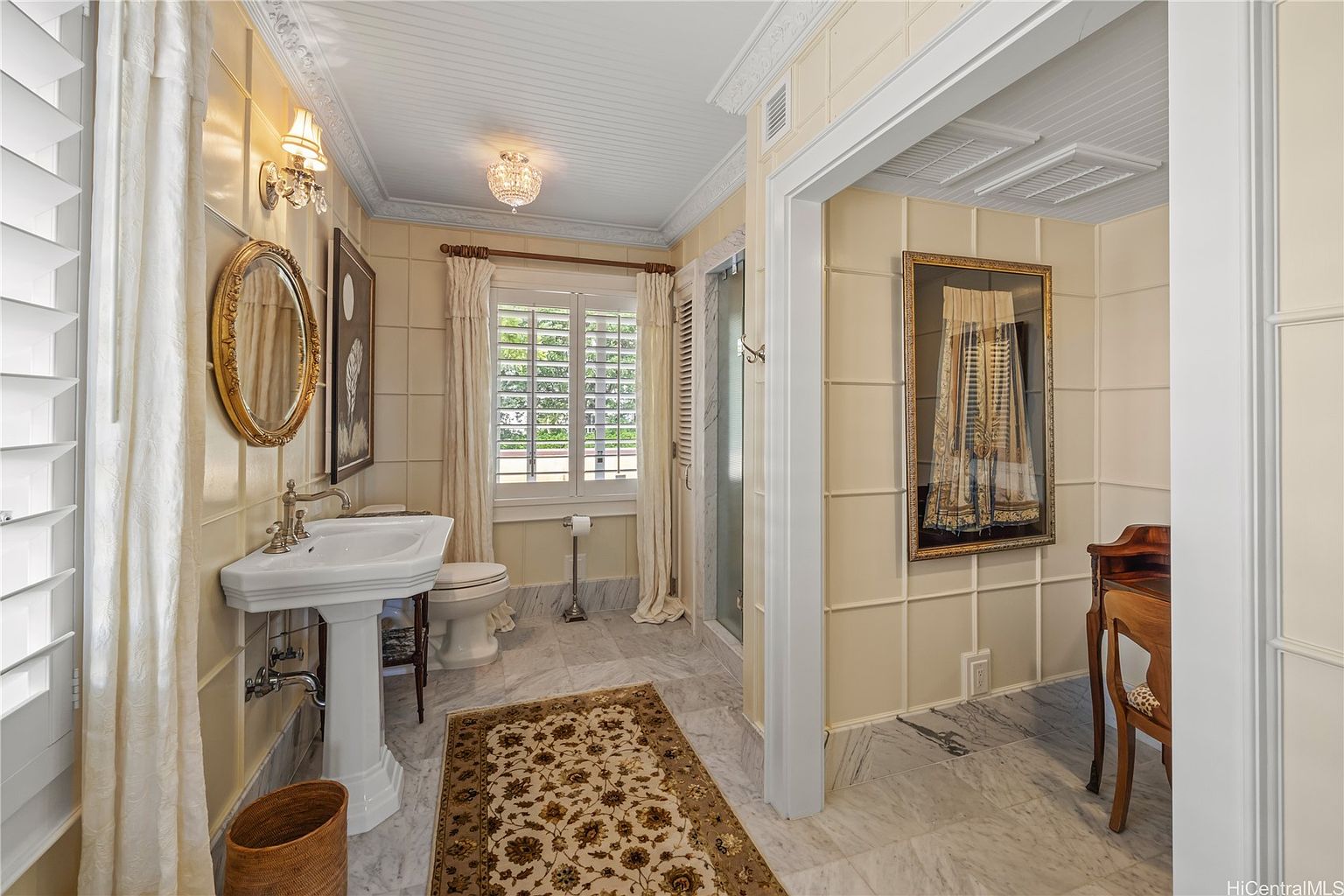 This is a well-appointed bathroom featuring a pedestal sink with a gold-framed oval mirror above it, a toilet, and a glass-enclosed shower. The walls are paneled in a light yellow tone, complemented by marble flooring and a decorative rug. The room exudes a classic and elegant style, enhanced by the chandelier and framed artwork.