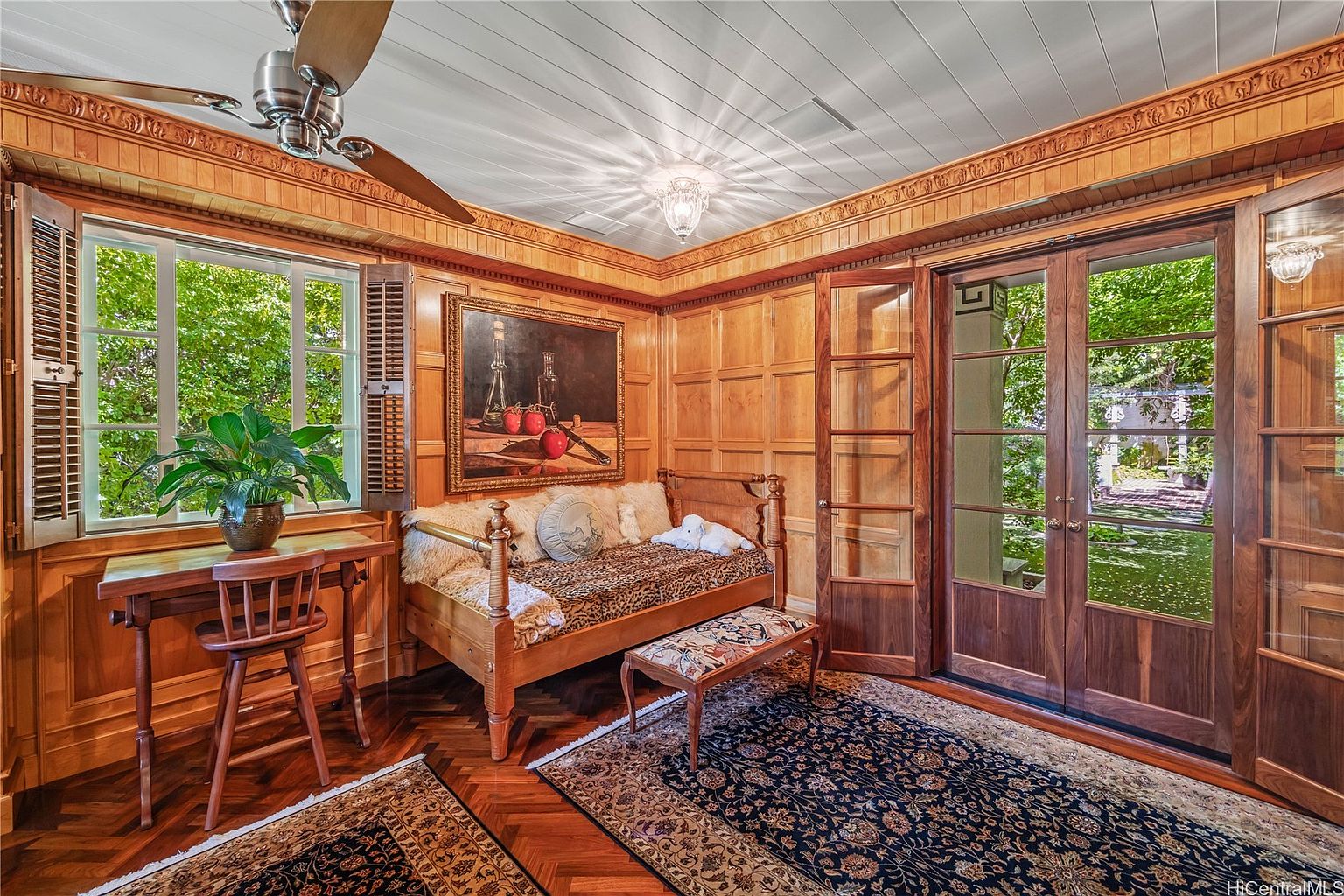 This sunroom features wood-paneled walls and ceiling, creating a warm and inviting atmosphere. A daybed with decorative pillows sits beneath a framed painting, while a desk and chair are positioned by the window, which is covered by shutters. The room is illuminated by a chandelier and natural light, and a patterned rug covers the hardwood floor.
