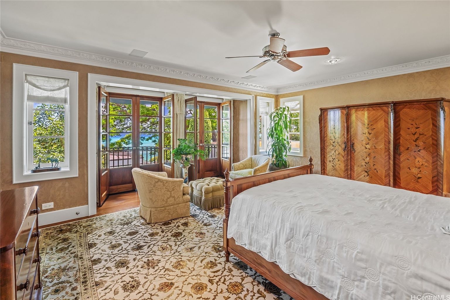 This primary bedroom features a large bed with white linens, a decorative rug, and a seating area near a set of French doors that lead to a balcony with an ocean view. The room is decorated in warm tones with detailed crown molding and a ceiling fan, creating a luxurious and inviting atmosphere. A large wooden wardrobe stands against the wall, adding to the room's elegance.