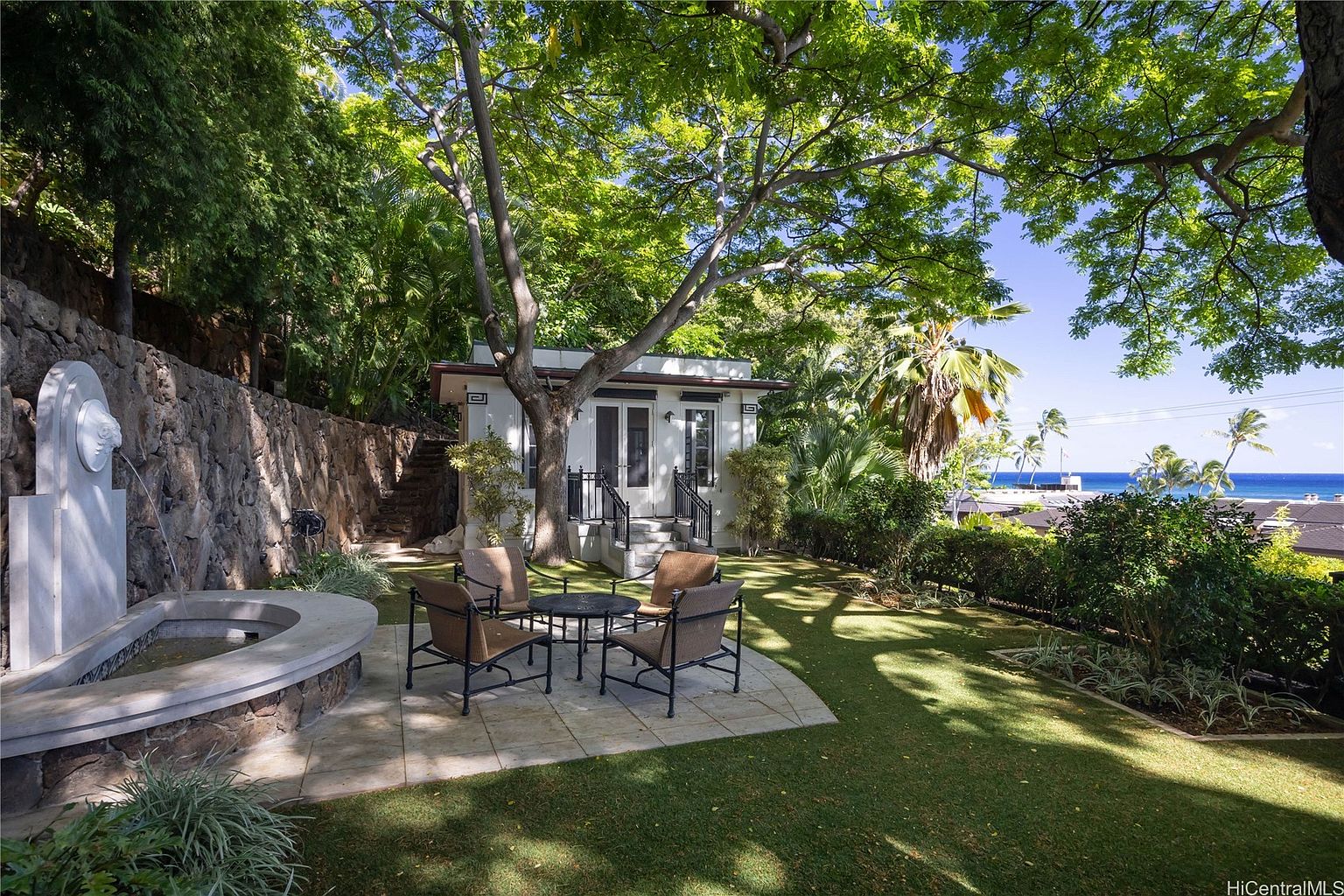 This image showcases a beautifully landscaped yard with a charming white cottage-style building in the background. A stone patio with outdoor seating provides a relaxing space, while lush greenery and mature trees create a serene and private atmosphere. In the distance, a glimpse of the ocean adds to the property's appeal.
