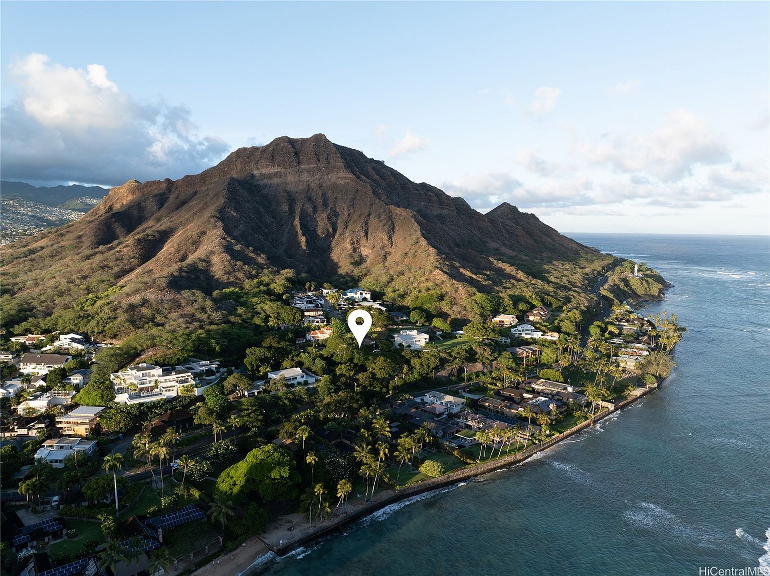 This aerial shot showcases a luxurious coastal property nestled against a mountain backdrop. The homes feature modern architectural styles with lush landscaping and palm trees lining the shoreline. The turquoise ocean provides a stunning contrast to the green foliage and the brown mountain, creating an idyllic and exclusive setting.