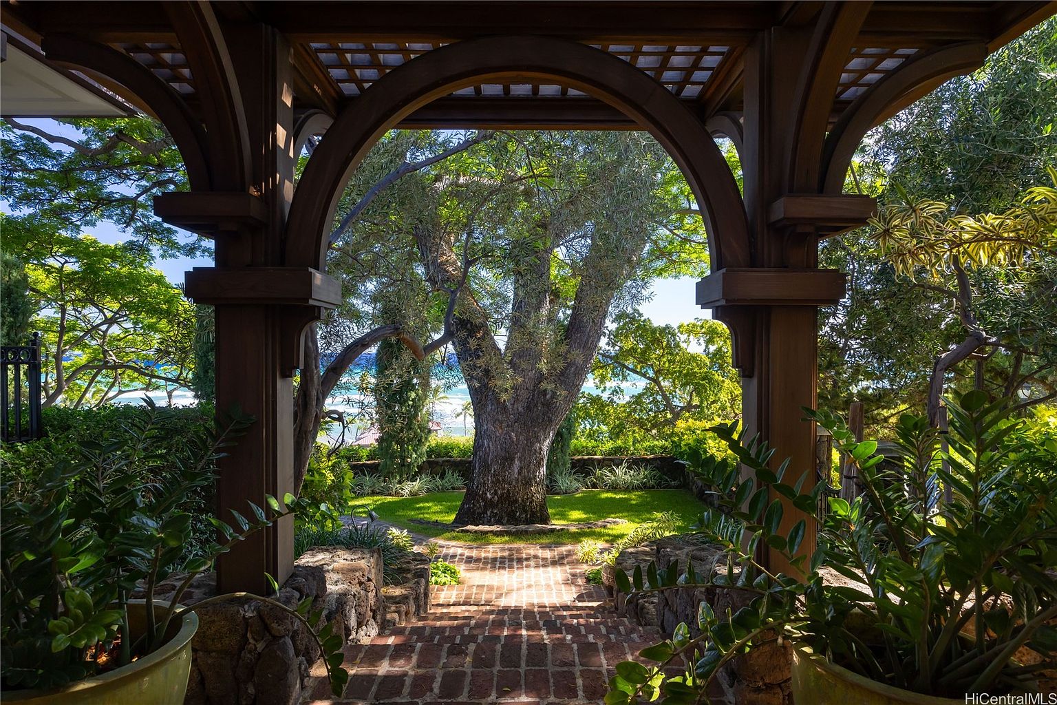 This image showcases a beautifully landscaped yard and garden, framed by a wooden pergola. A brick pathway leads through the lush greenery towards a large tree, with glimpses of the ocean visible in the background. The scene evokes a sense of tranquility and privacy, highlighting the property's outdoor living space.
