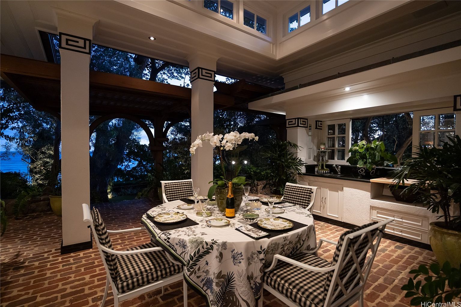 This image showcases an elegant outdoor dining area on a brick patio. A round table is set for a meal, adorned with a floral tablecloth and surrounded by chairs with black and white checkered cushions. The space is partially covered, with decorative columns and a view of lush greenery and a glimpse of the ocean in the background, creating a luxurious and inviting atmosphere.