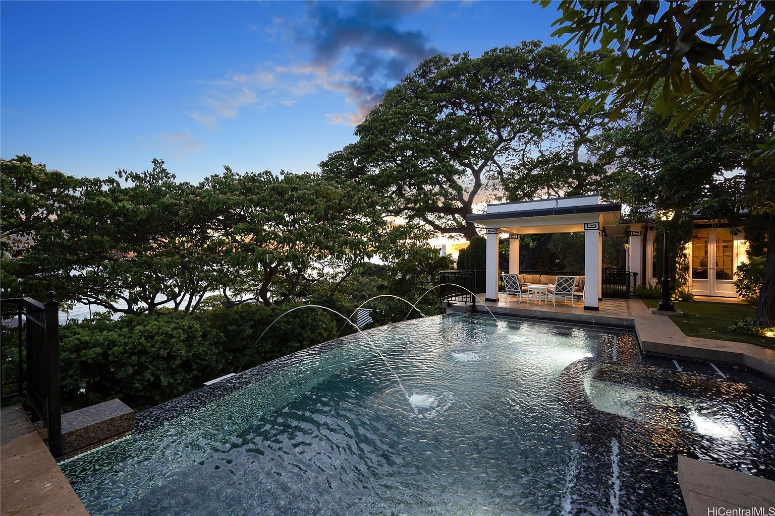 This image showcases a luxurious infinity pool with water features, set against a backdrop of lush greenery and a picturesque sunset sky. A covered pavilion with seating is situated adjacent to the pool, offering a relaxing space for entertaining. The overall impression is one of tranquility and high-end outdoor living.