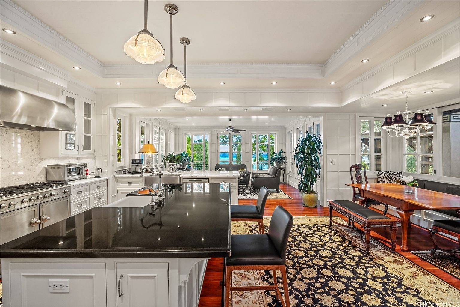 This is a bright and spacious kitchen that seamlessly transitions into a dining and living area. The kitchen features a large island with a black countertop, white cabinetry, and stainless steel appliances. The dining area includes a wooden table with bench seating, and the living area offers comfortable seating with a view of the ocean through large windows. The overall impression is one of luxury and comfort.