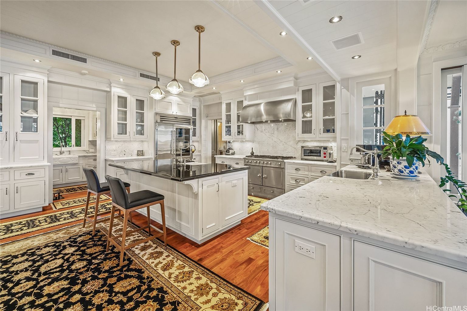This is a bright and spacious kitchen featuring white cabinetry, marble countertops and backsplash, and stainless steel appliances. A large island with a black countertop provides ample workspace and seating, while pendant lights add a touch of elegance. The kitchen is well-lit and appears to be designed for both functionality and style, with a warm wood floor and decorative rug adding character.