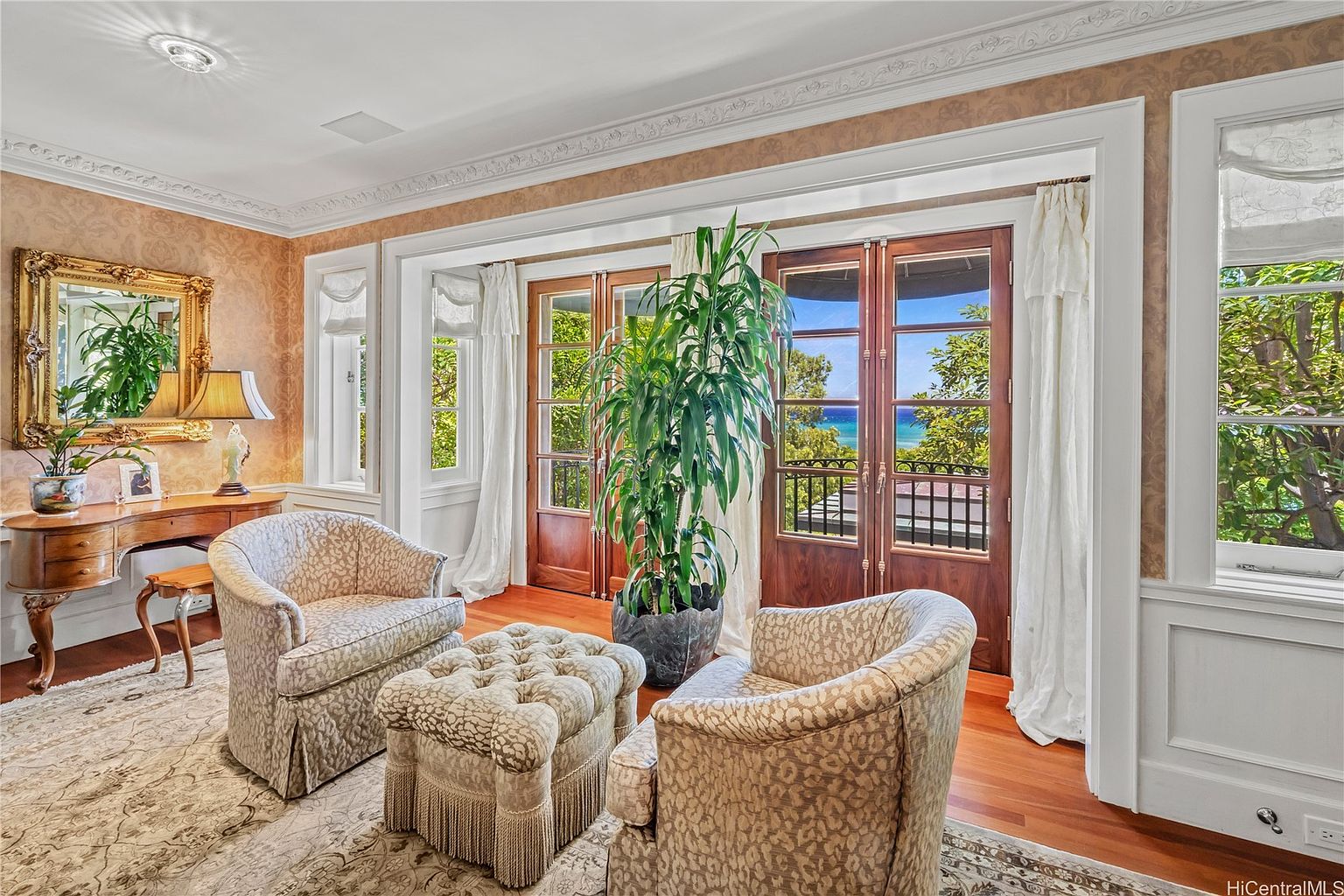 This is an elegant living room featuring two patterned armchairs and a tufted ottoman, creating a cozy seating area. A wooden desk with a gold-framed mirror sits against the wall, complemented by a table lamp. Large wooden doors lead to a balcony with ocean views, enhancing the room's appeal and natural light.
