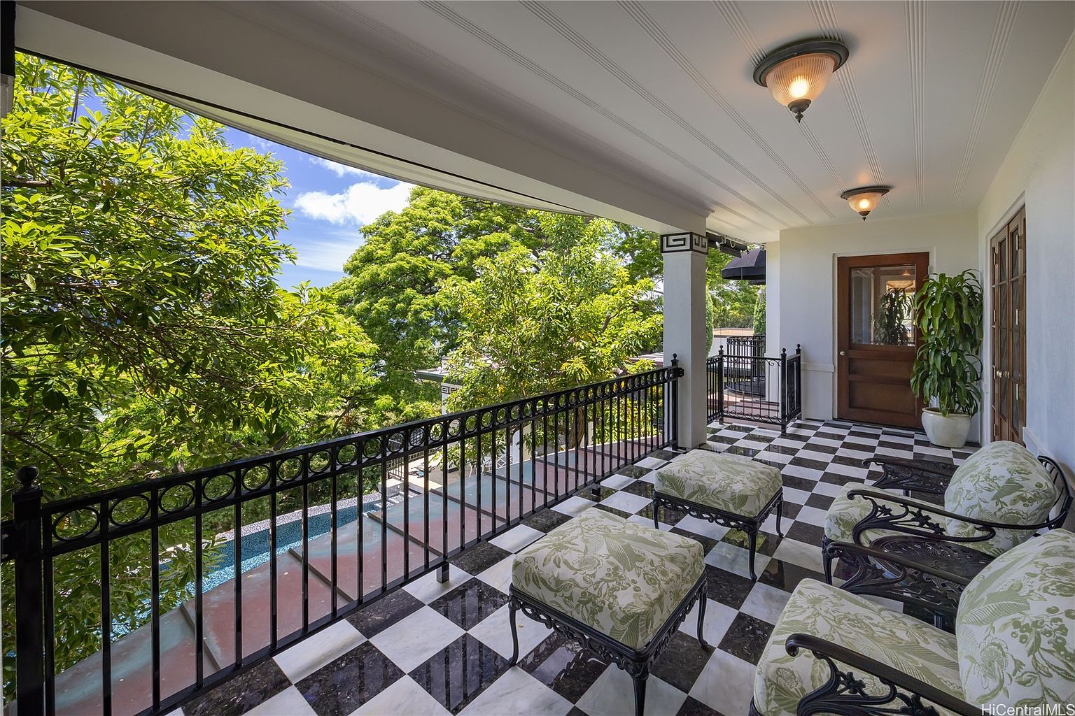 This image showcases a stylish balcony with a black and white checkered floor, ornate black railings, and comfortable seating arrangements including chairs and ottomans with floral patterns. The balcony offers a view of lush green trees and a glimpse of a pool below, creating a serene and inviting outdoor living space. The ceiling is white with recessed lighting.