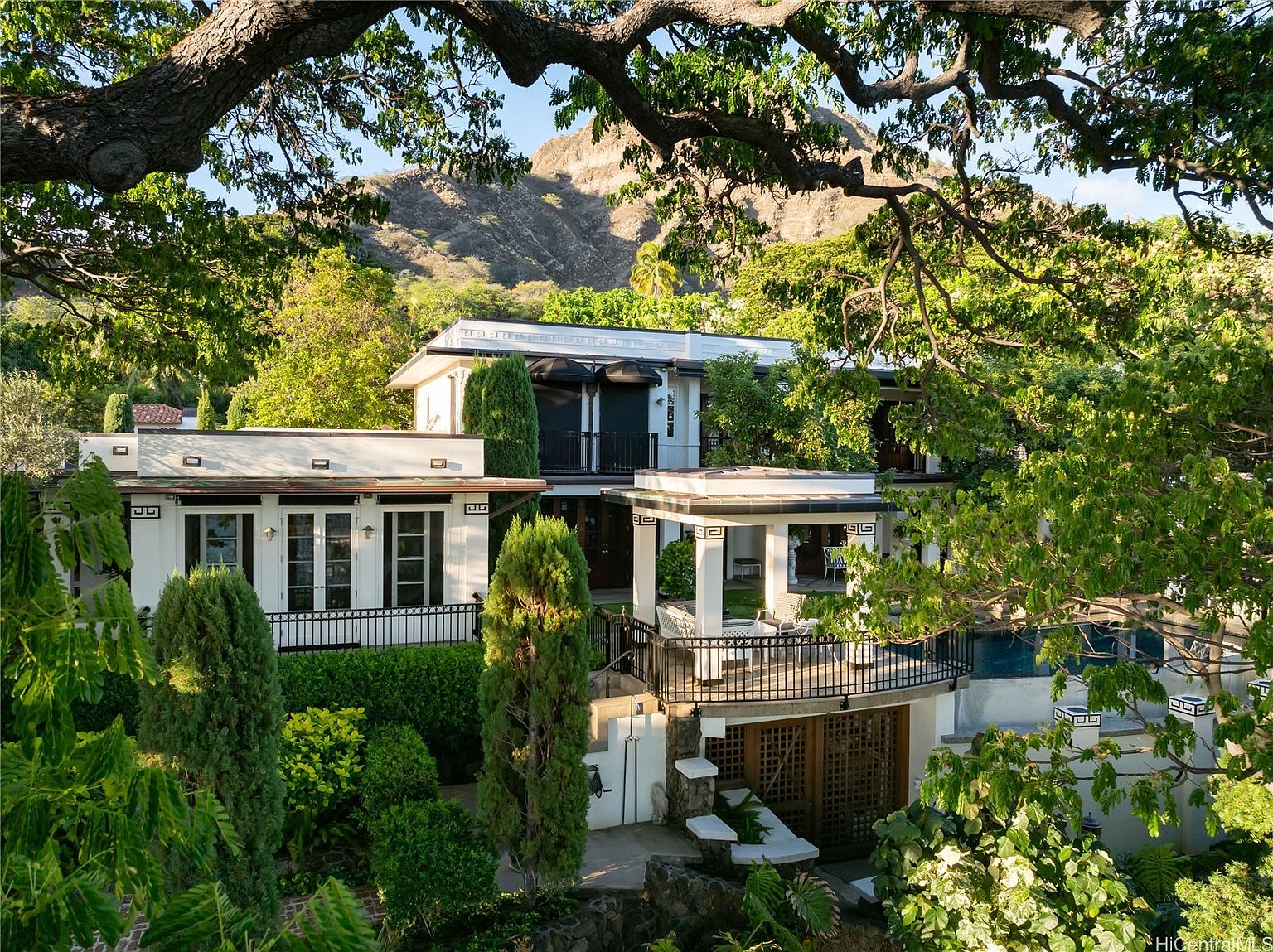 This image showcases the front exterior of a multi-level home, framed by lush greenery and a large tree branch overhead. The architecture features a blend of modern and classic elements, with white walls, dark window frames, and decorative Greek key patterns. A balcony and patio area add to the home's appeal, while a mountain is visible in the background, enhancing the property's scenic setting.