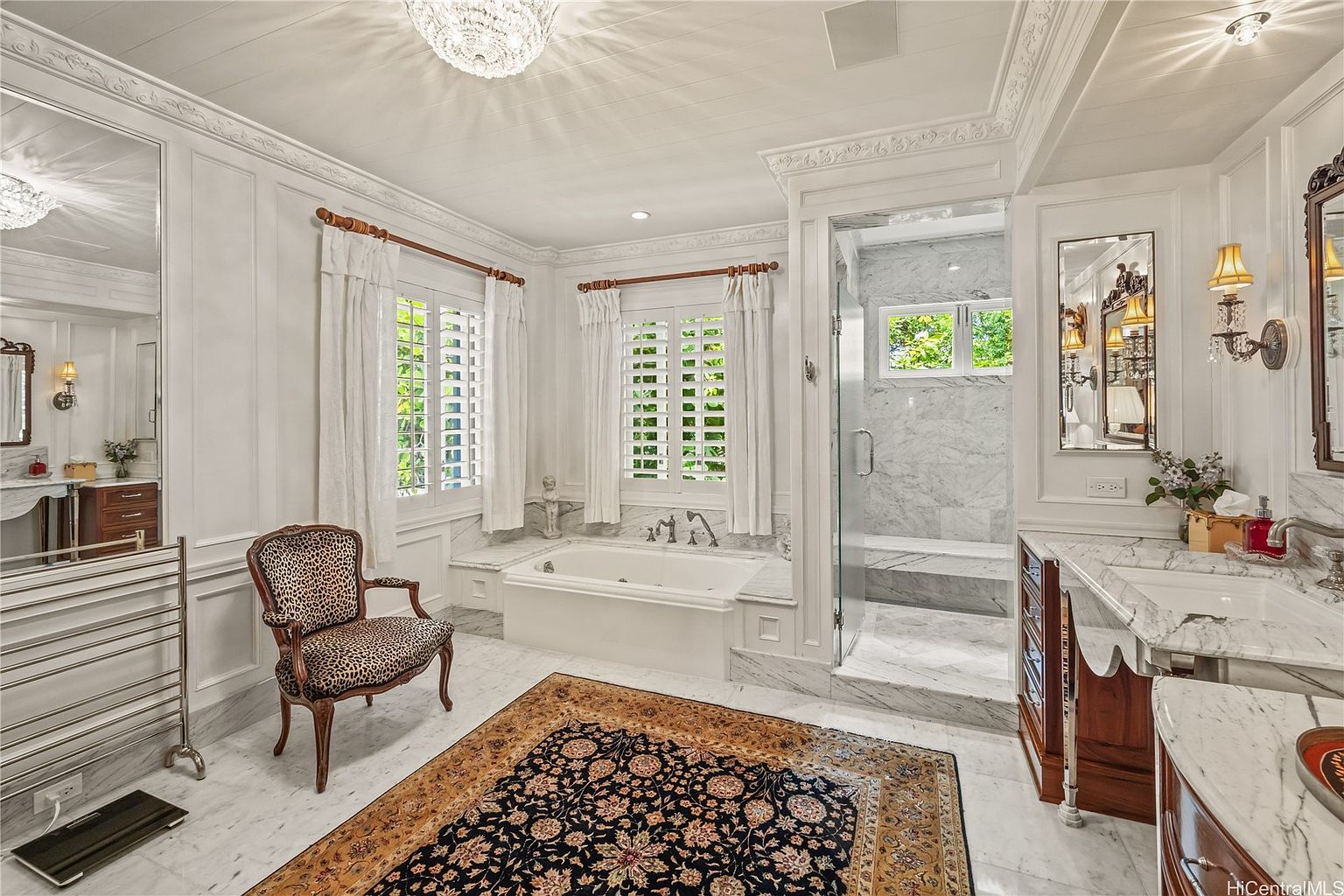 This is a luxurious primary bathroom featuring a soaking tub, a glass-enclosed shower, and dual vanities with marble countertops. The room is decorated in a classic style with white walls, detailed molding, and a chandelier. A patterned rug adds warmth and character to the marble flooring.