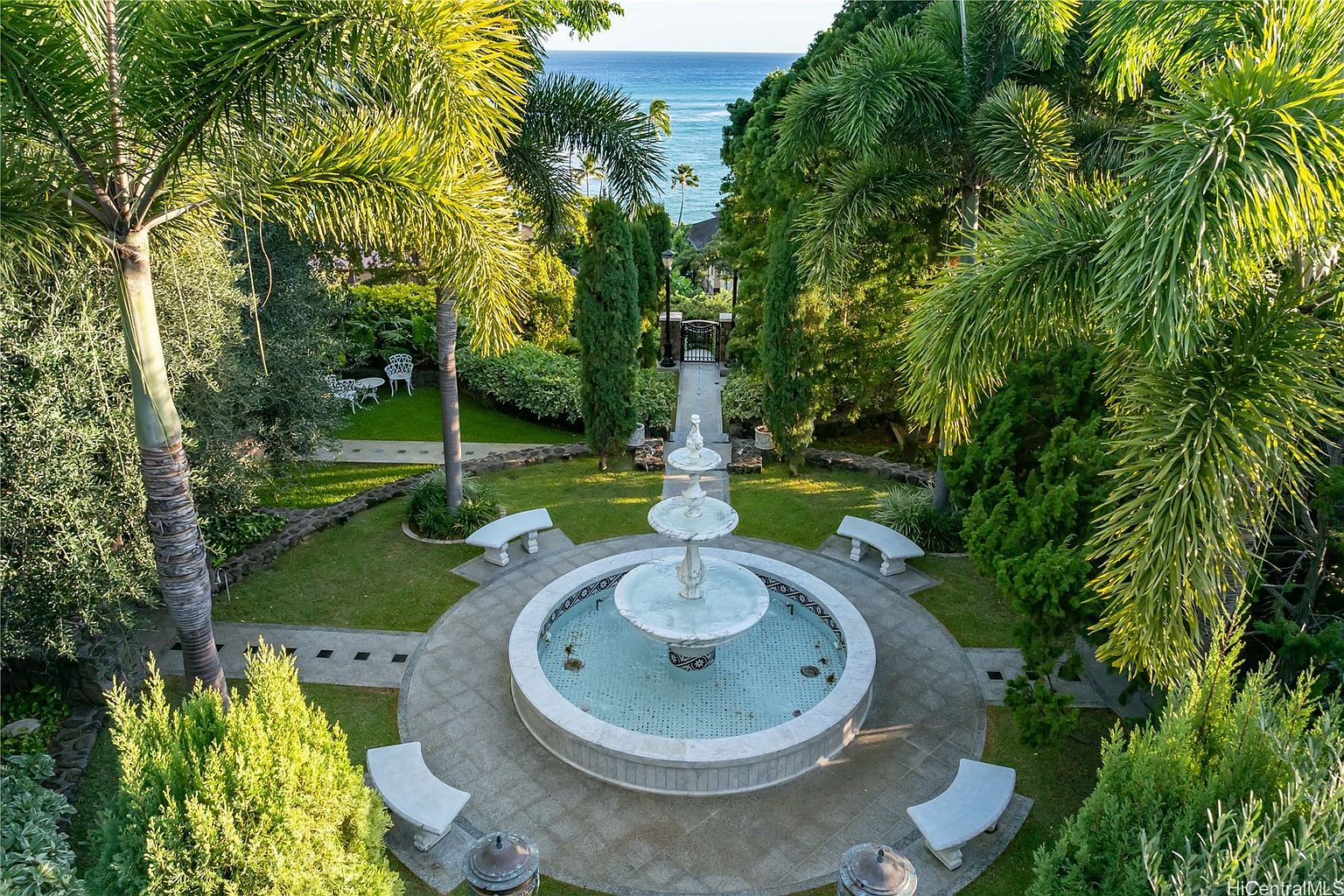 This aerial view showcases a meticulously landscaped garden featuring a tiered fountain as its centerpiece. Stone benches surround the fountain, inviting relaxation amidst lush greenery and palm trees. A pathway leads towards a gated entrance, offering a glimpse of the ocean in the distance, creating a serene and luxurious outdoor space.