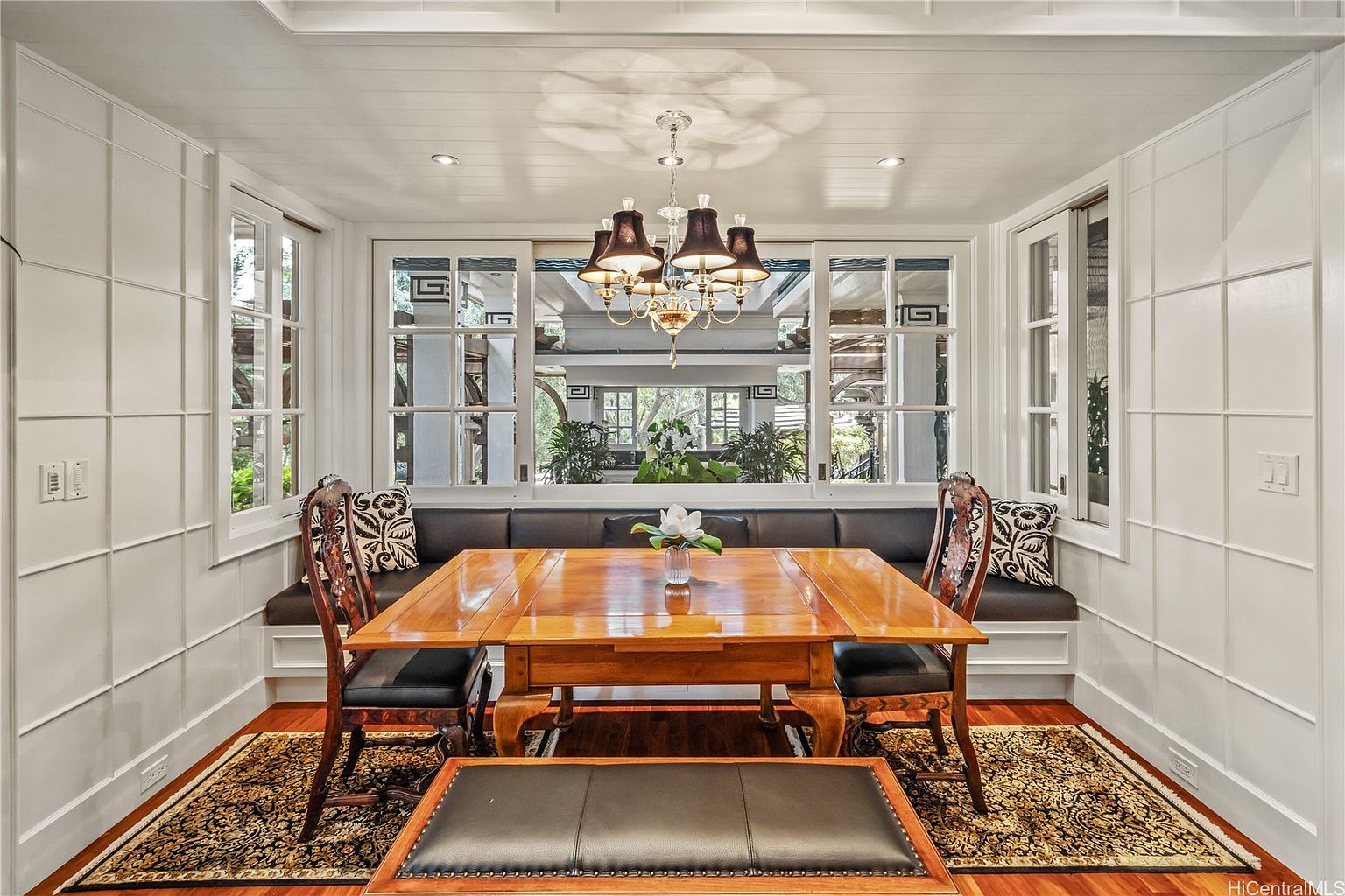 This is an interior shot of a dining room featuring a wooden dining table with chairs and a bench, set on a patterned rug. A built-in banquette seating area is positioned against a large window, providing natural light. The room is decorated in a classic style with white paneled walls and a chandelier overhead, creating an elegant and inviting atmosphere.