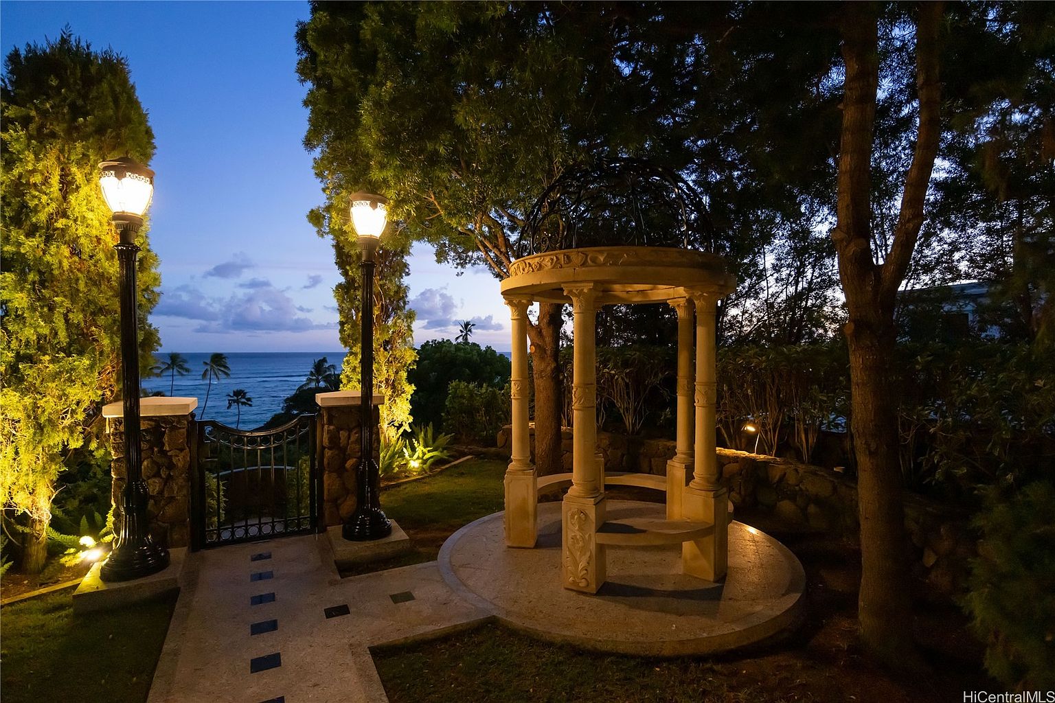 This image showcases a beautifully landscaped yard or garden at dusk, featuring a charming gazebo with stone columns and a circular base. A pathway leads to a gate with a view of the ocean and palm trees in the distance, flanked by elegant lampposts. The scene is illuminated by soft lighting, creating a serene and inviting atmosphere.
