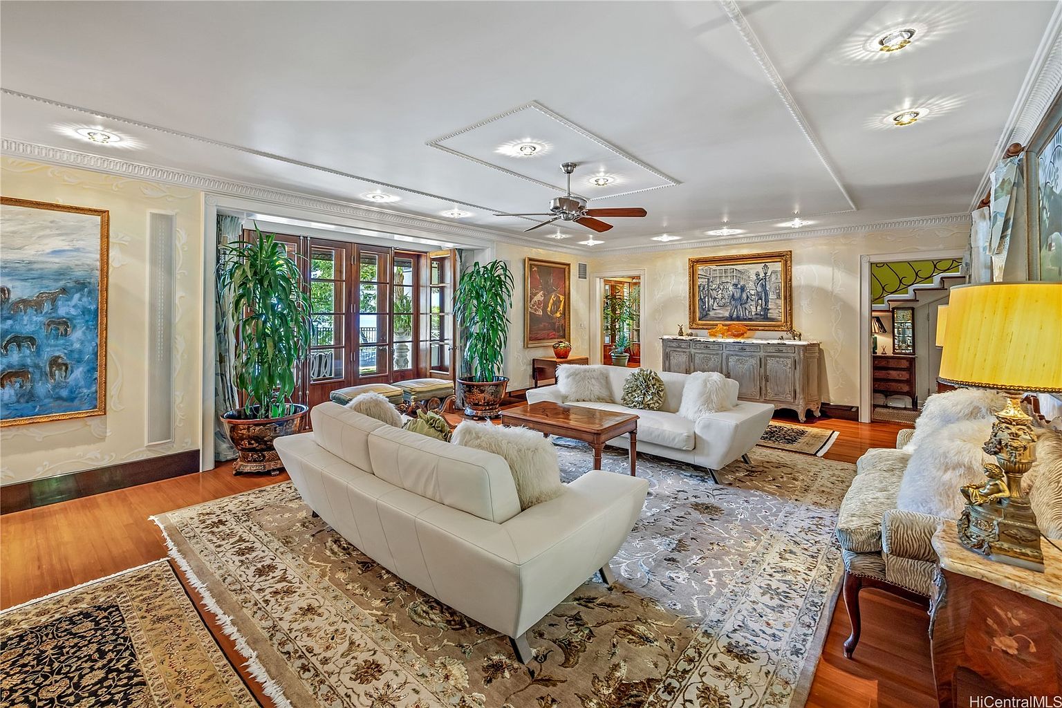 This is an interior shot of a luxurious living room featuring two white sofas, a wooden coffee table, and an ornate area rug. The room is well-lit with recessed lighting and natural light from the large doors, complemented by artwork and potted plants. The overall impression is one of elegance and comfort.